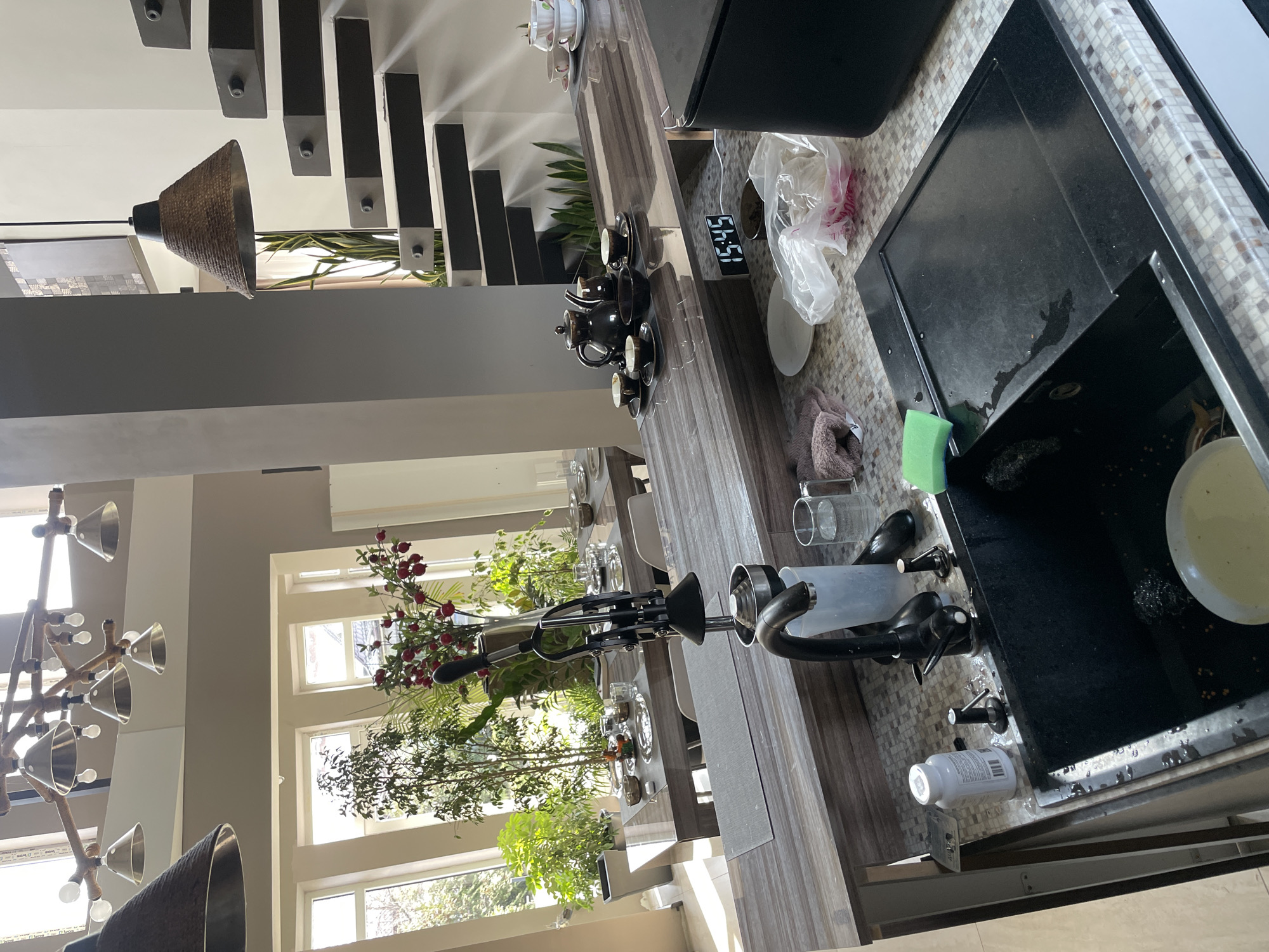 The image shows a portion of a kitchen in an unknown location. The scene includes a sink filled with dirty dishes, a faucet, and various cleaning supplies. A digital clock reads 15:45. Adjacent to the sink is a countertop with a tea set, cups and saucers, and a plastic shopping bag. A dining table is set in the background, decorated with flowers and plants in front of a window. Part of a modern staircase with a black railing is visible in the background. The room has a modern interior design with unique lighting fixtures. No people are visible in the scene.