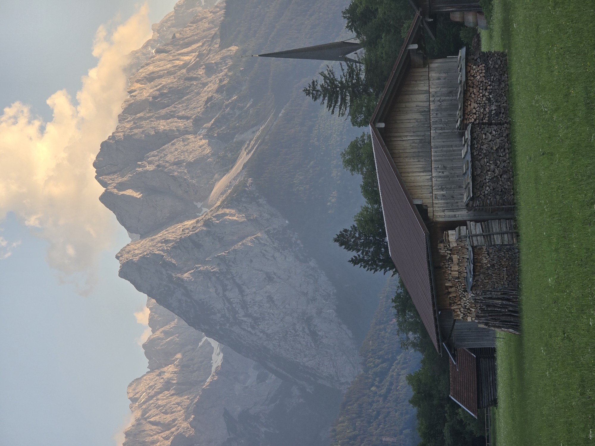 The image depicts a serene mountain landscape near Garmisch-Partenkirchen, Germany, bathed in the soft, warm light of late afternoon or early evening. In the foreground, a weathered wooden barn stands prominently, its walls constructed of vertical planks and its roof covered in dark shingles. Large stacks of neatly cut firewood are piled against the front and side of the barn, some covered with flat wooden tops. To the left, a smaller, similarly constructed wooden shed with a red-tiled roof is visible.
Behind the barn, a lush green meadow dotted with small white wildflowers stretches out. Further back, a dense forest of dark green coniferous trees forms a natural backdrop. Rising majestically behind the trees is a formidable mountain range, its craggy peaks illuminated by the setting sun, casting long shadows. The mountains exhibit a rugged texture with patches of snow visible on some of the higher elevations.
To the right of the frame, the slender, dark spire of a church steeple pierces the sky above the treeline, adding a touch of architectural interest to the natural scenery. The sky above is a pale blue, with soft, wispy clouds tinged with the golden hues of the sunset. The overall atmosphere is one of rural tranquility and natural grandeur.