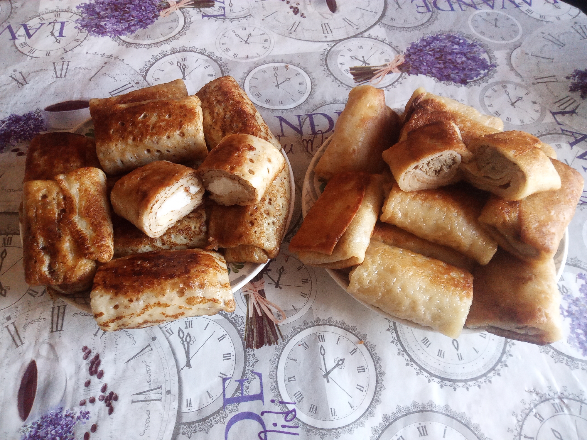 A top-down shot displays two plates of savory crepes, or blini, filled and rolled, arranged on a tablecloth adorned with vintage clock faces and sprigs of lavender. The crepes are golden brown and appear to be filled with a light-colored, creamy filling on the left, and a darker, meat-like filling on the right, visible in the cut sections of some crepes. The tablecloth features Roman numerals and decorative clock hands, suggesting a theme of time or antique aesthetics. The overall presentation is casual, possibly a home setting, with no individuals or discernible city landmarks from Usman, Russia, present in the frame.