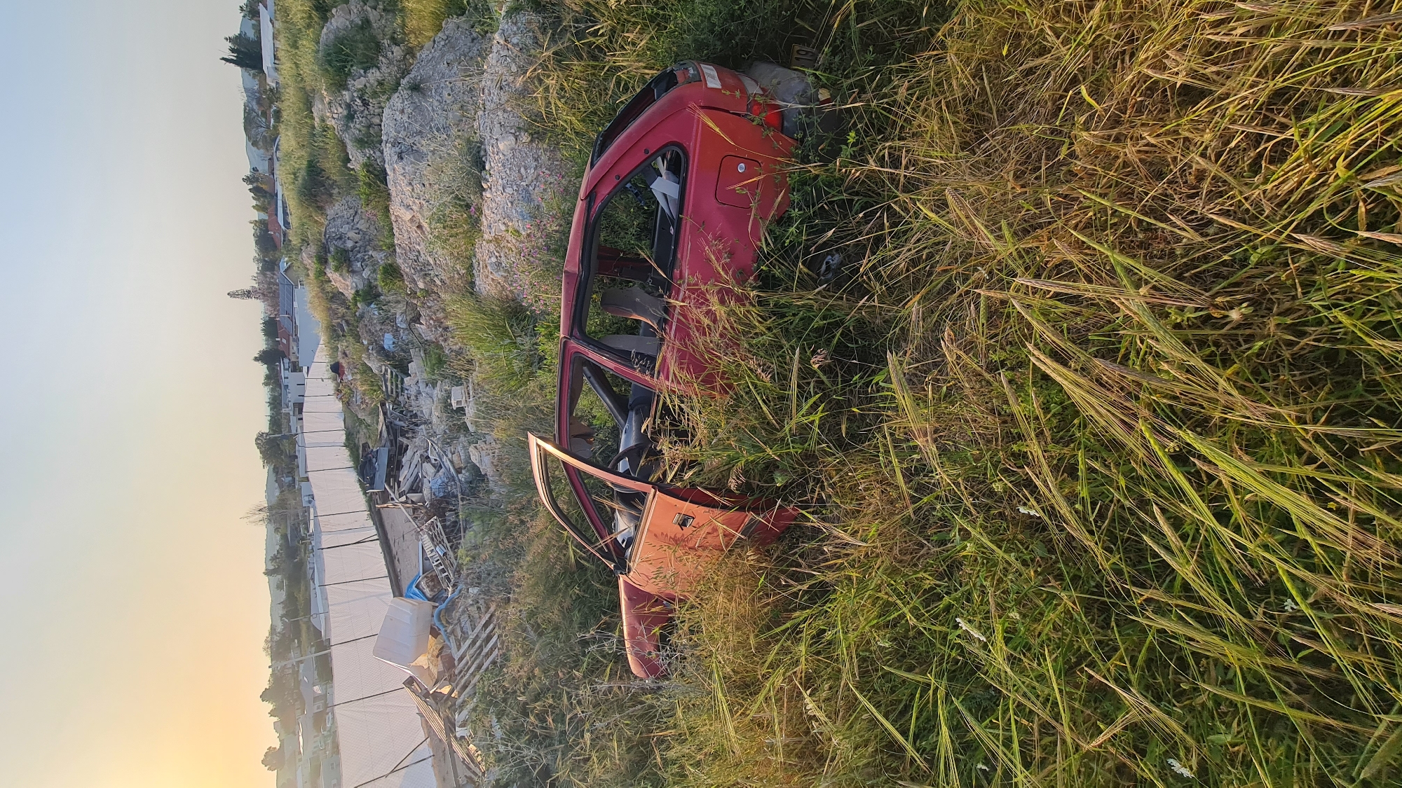 The image shows the rusted frame of a red car in Lachish Regional Council, Israel. The car is missing most of its body panels and the interior is visible. It rests in a field of tall, dry grass. In the background, there is a rocky hillside with scattered vegetation and some buildings. The sky is hazy. Debris and construction material are scattered nearby.
