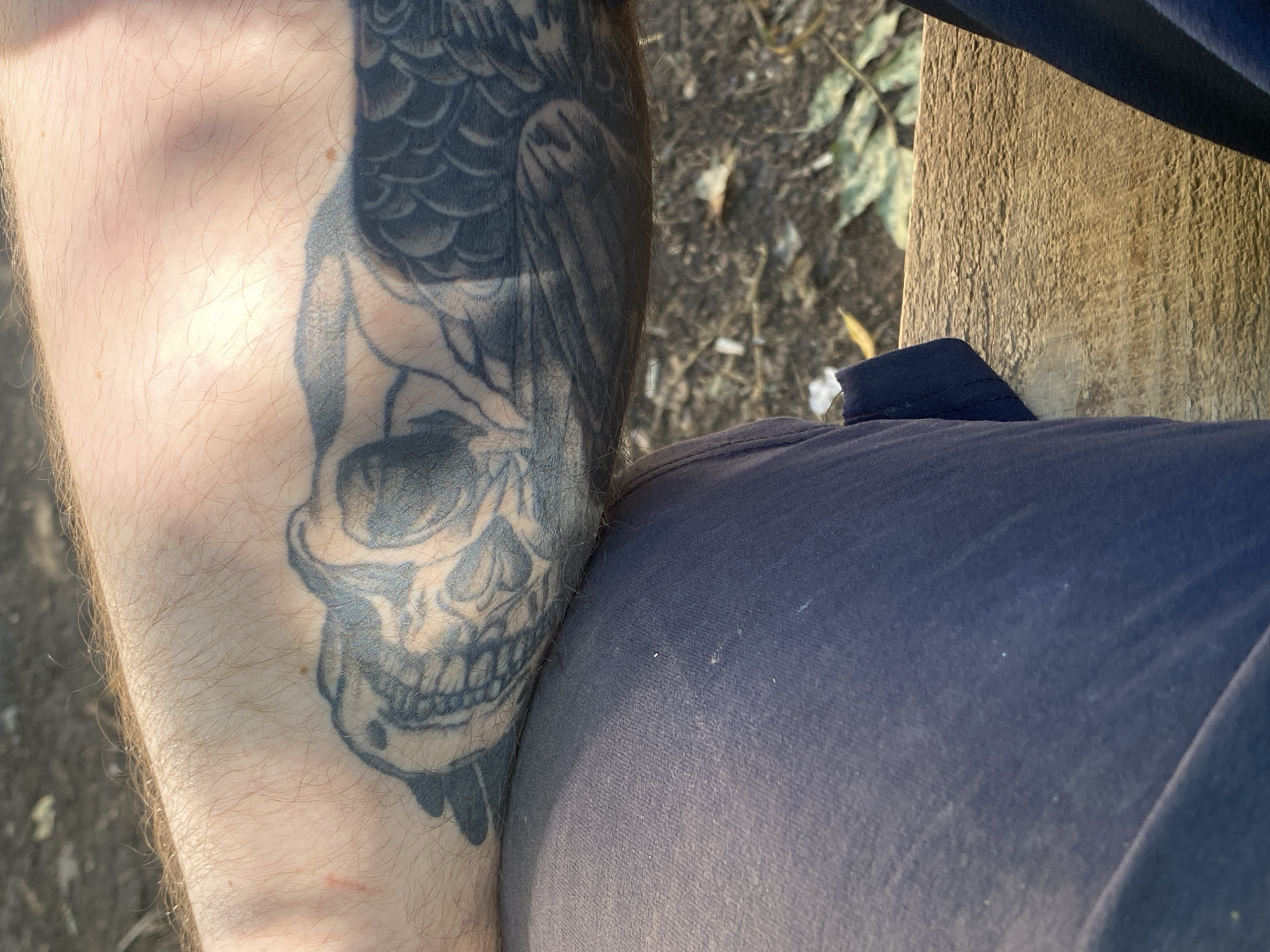 The image shows a close-up view of a person's arm with a tattoo and part of their clothing. The tattoo appears to be a black and gray depiction of a creature, possibly a dragon or a mythical bird, with intricate scales and wings. The skin is lightly tanned and has some visible hair. The person is wearing dark blue pants. The background is out of focus, showing a mix of dirt, leaves, and a wooden surface, suggesting an outdoor setting. The lighting indicates it is daytime, with some shadows cast on the arm. The location is unknown.
