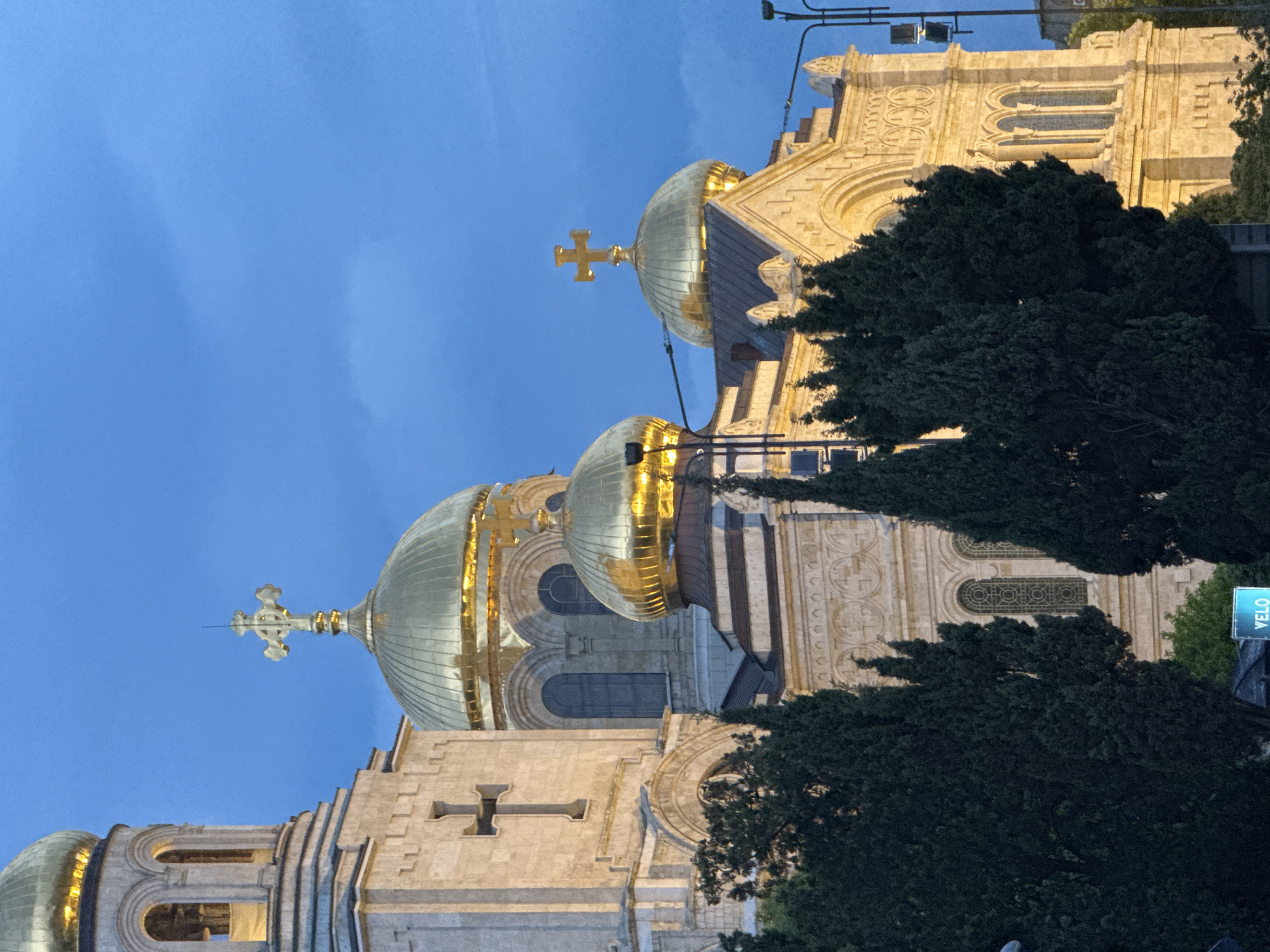The image shows the Dormition of the Mother of God Cathedral in Varna, Bulgaria, at dusk or dawn. The cathedral, constructed of light-colored stone, features several golden domes topped with crosses. The sky is a deep blue, suggesting twilight. Parts of the building are illuminated with artificial light, highlighting architectural details. Trees partially obscure the base of the building on the right. A "VELO" sign can be seen on the bottom right.