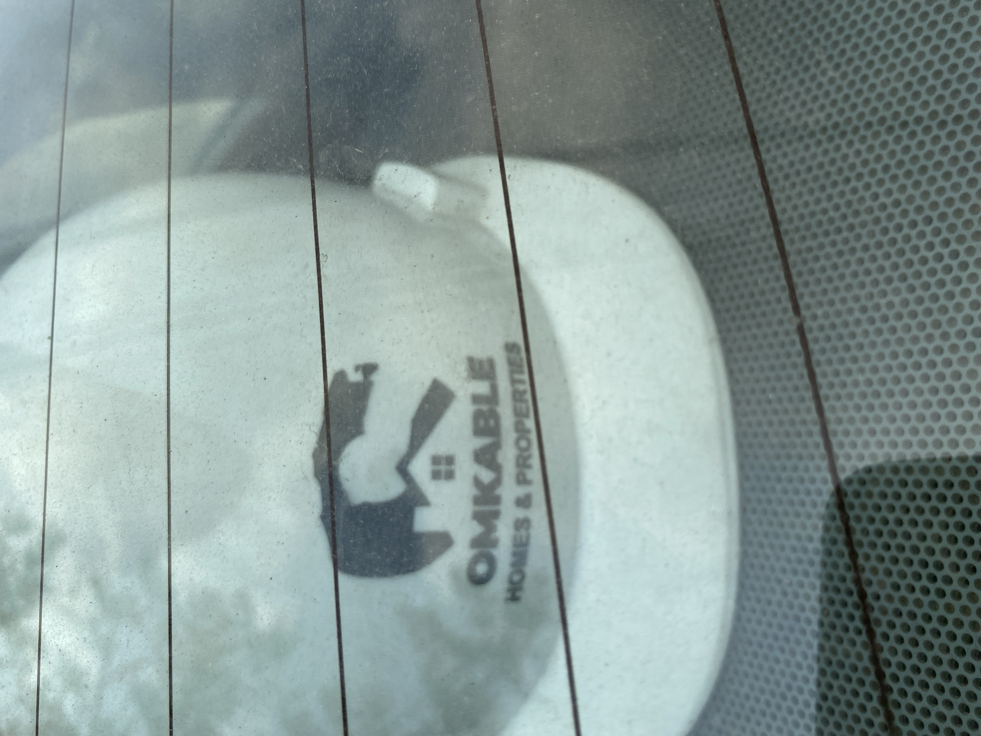 The image captures a white hard hat seen through what appears to be a slightly dirty or dusty car window, possibly a rear windshield, characterized by prominent vertical defroster lines. The hard hat is positioned within the vehicle, resting on a surface that is out of focus. To the right, a dark, perforated pattern is visible, suggesting a part of the car's interior like a speaker grille or a textured panel.
A black logo and text are clearly visible on the hard hat. The logo is a stylized depiction of a house or building. Adjacent to it, the word "OMKABLE" is printed in large, bold letters, with "HOMES & PROPERTIES" written beneath it in a smaller font. This visible text indicates an affiliation with a real estate or construction company named Omkable Homes & Properties.
The scene is illuminated by daylight, suggesting the photograph was taken during the day. There are no individuals present, and no specific activity is taking place. The image focuses on the stationary hard hat within the vehicle, potentially in Lagos, Nigeria, given the contextual information.