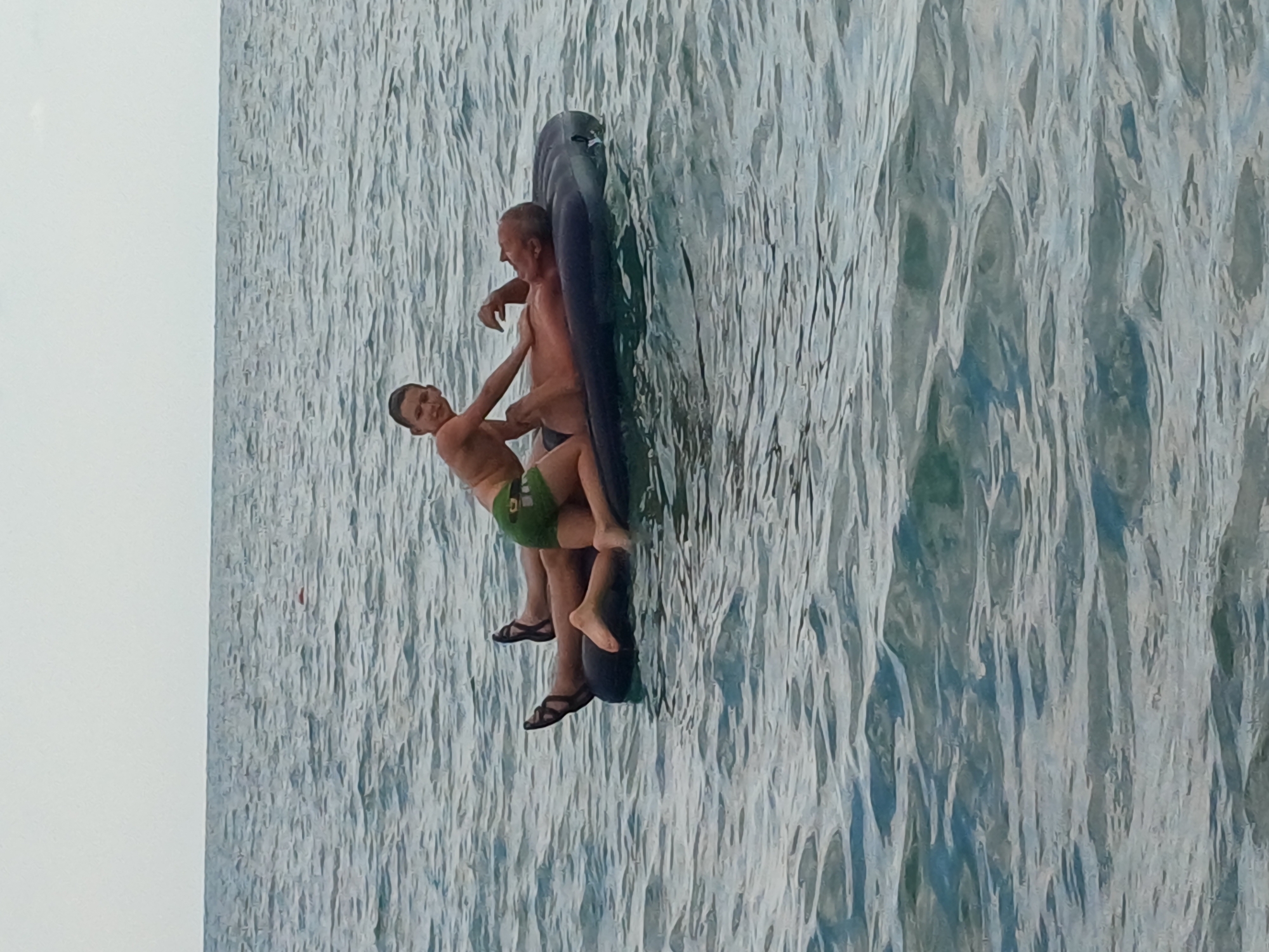 The image shows two people, an adult male and a young boy, on an inflatable mattress in the water. The adult male is supporting the boy, who is hanging onto him. Both are wearing swimwear. The boy is wearing green swim shorts, and the adult male appears to be wearing dark swim trunks. They are in a body of water that appears to be the sea, with a textured surface indicating ripples or waves. The lighting suggests it is daytime. The scene takes place outdoors. There is no visible text in the image.