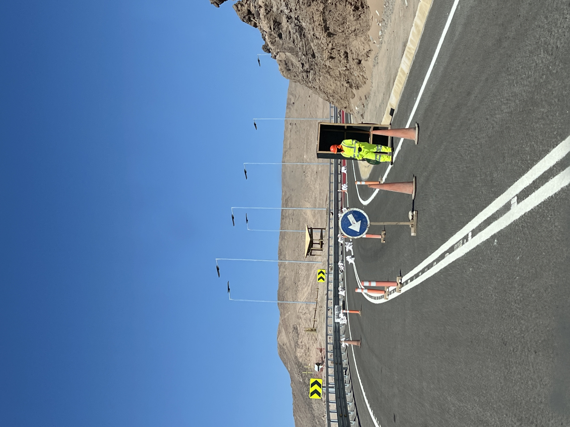 A photograph depicts a highway scene near Arica, Chile. A road worker in high-visibility clothing stands within a fenced construction zone, flanked by traffic cones. Roadside infrastructure, including chevron signs and lane markers, is visible. Overhead wires extend from poles along the road, against a backdrop of arid, mountainous terrain. The sky is clear and blue.