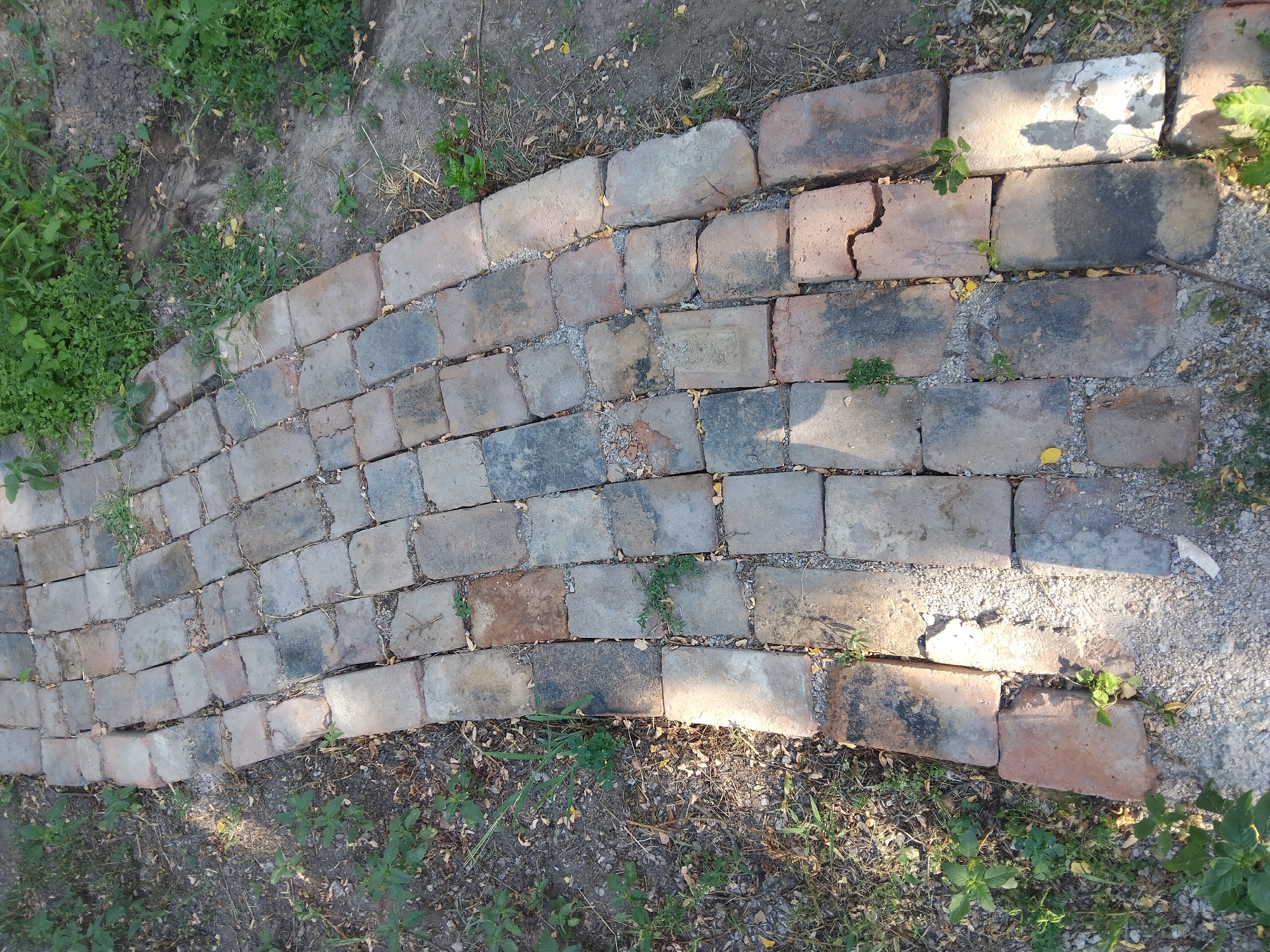 A winding brick pathway, constructed from weathered, rectangular bricks of varying shades of red, gray, and black, traverses a dusty, unpaved area. The bricks are laid in a staggered pattern, creating a slightly uneven surface. Green vegetation, including grass and small leafy plants, grows sporadically along the edges of the path and in the gaps between the bricks, suggesting an outdoor, possibly neglected, environment. The ground surrounding the path is composed of dirt, small stones, and dried leaves, indicating a natural or semi-natural setting. The lighting suggests it is daytime, with some shadows cast across the pathway. This scene is located in Pervozvanivka, Ukraine.