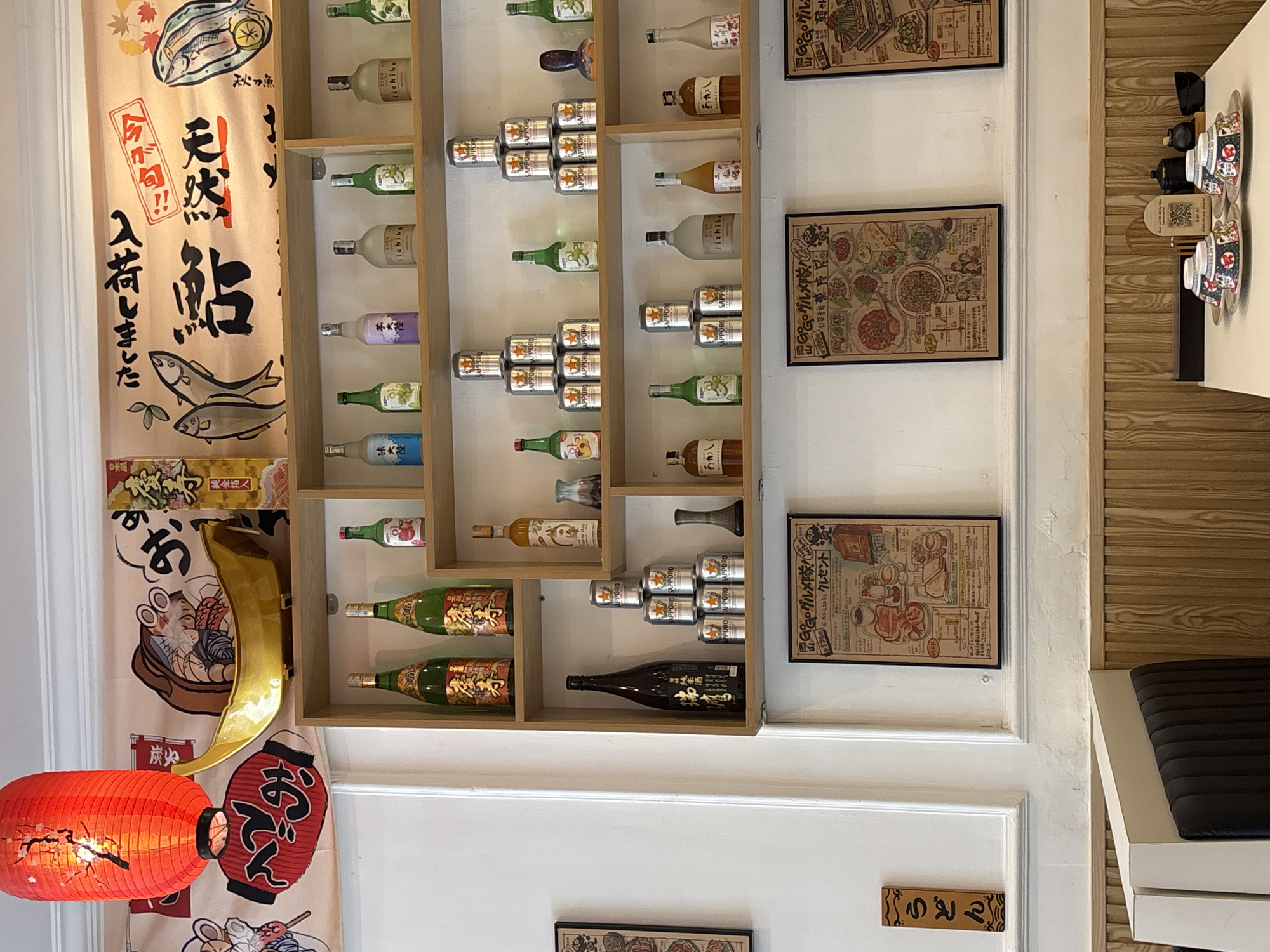 The image shows an indoor wall decorated with Japanese-themed items.
On the left is a decorative fabric panel featuring Japanese writing, illustrations of fish, a bowl of noodles, and a red paper lantern. Adjacent to the panel is a wooden shelf containing various bottles and stacked cans of Sapporo beer. Three framed posters are arranged vertically on the right wall, each depicting illustrated Japanese dishes and menus with Japanese writing. The wall is white with a decorative border.
On the right, part of a light-wood patterned wall can be seen, with small ceramic teacups stacked neatly. A portion of a bench with a black cushion is visible at the lower right. A small wooden sign with Japanese characters is located at the bottom center of the wall.
There are no visible people or interactions in this scene.
Based on the Japanese décor and style, the image likely originates from a Japanese restaurant or establishment. No specific city is indicated.