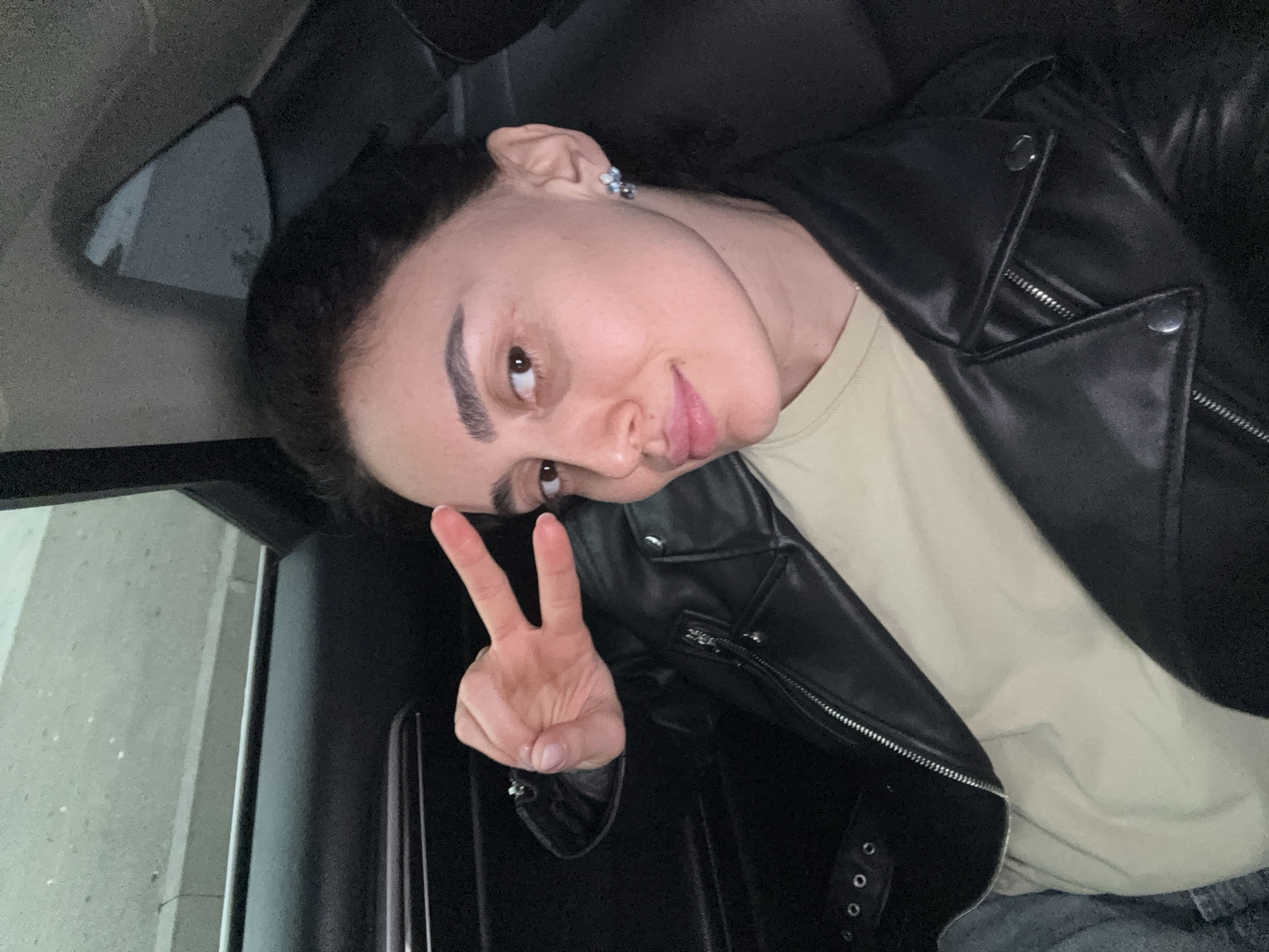 The scene is set inside the rear passenger compartment of a vehicle in Tbilisi, Georgia. A female individual, appearing to be in her early to mid-20s, occupies the frame, looking directly into the camera with a slight smile. She has dark hair, likely pulled back, and is wearing a black leather jacket over a plain light beige or cream-colored t-shirt. Blue denim fabric is partially visible below the jacket. Her left ear displays a small, multi-faceted stud earring. Her right hand is raised, performing a "peace sign" (V-gesture) with her index and middle fingers extended. The car's interior features dark trim on the door and window frame. The exterior view through the window is blurry, showing indistinct grey urban structures and ambient light, preventing identification of a precise location within Tbilisi.