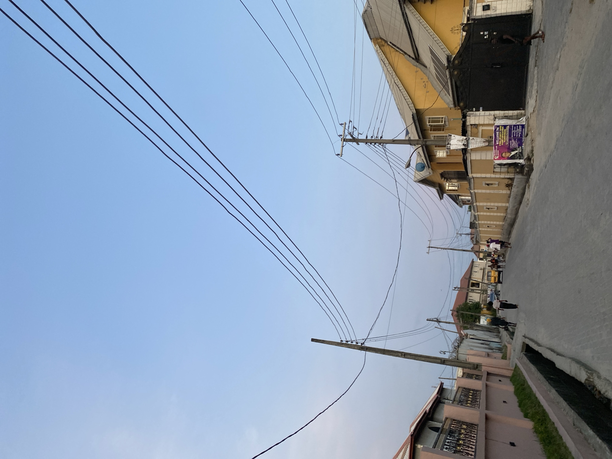 This image depicts a residential street scene in Iranla, Nigeria, under a clear, light blue sky. Overhead, numerous black electrical wires traverse the frame, with four parallel lines prominently visible in the upper right quadrant. The street surface is paved with gray asphalt or concrete, showing some wear.
On the left side of the street, an open, rectangular concrete drainage channel runs parallel to the roadway. Beyond this channel, a low fence with a pinkish-peach wall and black ornate metal railings borders properties. Portions of a building with a light-colored facade and a dark roof are visible behind this fence. Further down the left side, additional houses with various architectural styles and roofing are present. A wooden utility pole stands on the left.
On the right side of the street, a large, two-story building with bright yellow exterior walls and a gray corrugated metal roof is prominent. This building features multiple windows, some open, and solar panels are visible on its roof. A tall black metal gate serves as an entrance to this property. Adjacent to the gate, a purple and white banner advertises a "WELCOME TO FIRE, PURITY AND POWER CONFERENCE," displaying an image of a man in a suit. A concrete utility pole with multiple cross-arms and insulators supports numerous electrical wires in front of this building. Another utility pole is further down the street on the right.
Several individuals are present on the street. In the foreground on the right, a person dressed in a dark shirt and dark shorts, wearing sandals, walks away from the camera. Further down on the left, two individuals walk away from the camera, one in a light-colored top and another in a dark top. On the right side, in the mid-ground, a yellow mini-bus or commercial vehicle is parked or in motion, with a group of approximately 5-7 people standing or walking near it. The scene appears to be late afternoon or early evening due to the quality of light.
