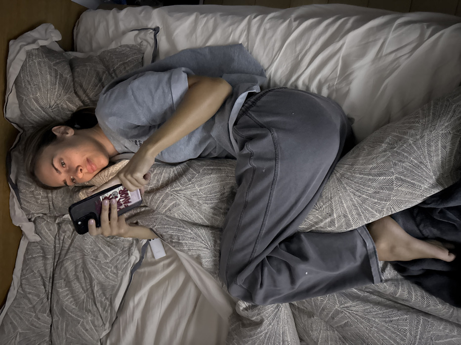 A person in gray loungewear lies sideways on a bed. The person's head rests on a pillow patterned with fern designs, while their body is angled across a white duvet. The person holds a phone with a decorated case in their hands, looking towards the camera. Bedding is crumpled around the individual. The overall light is subdued, giving the impression of relaxation or rest.