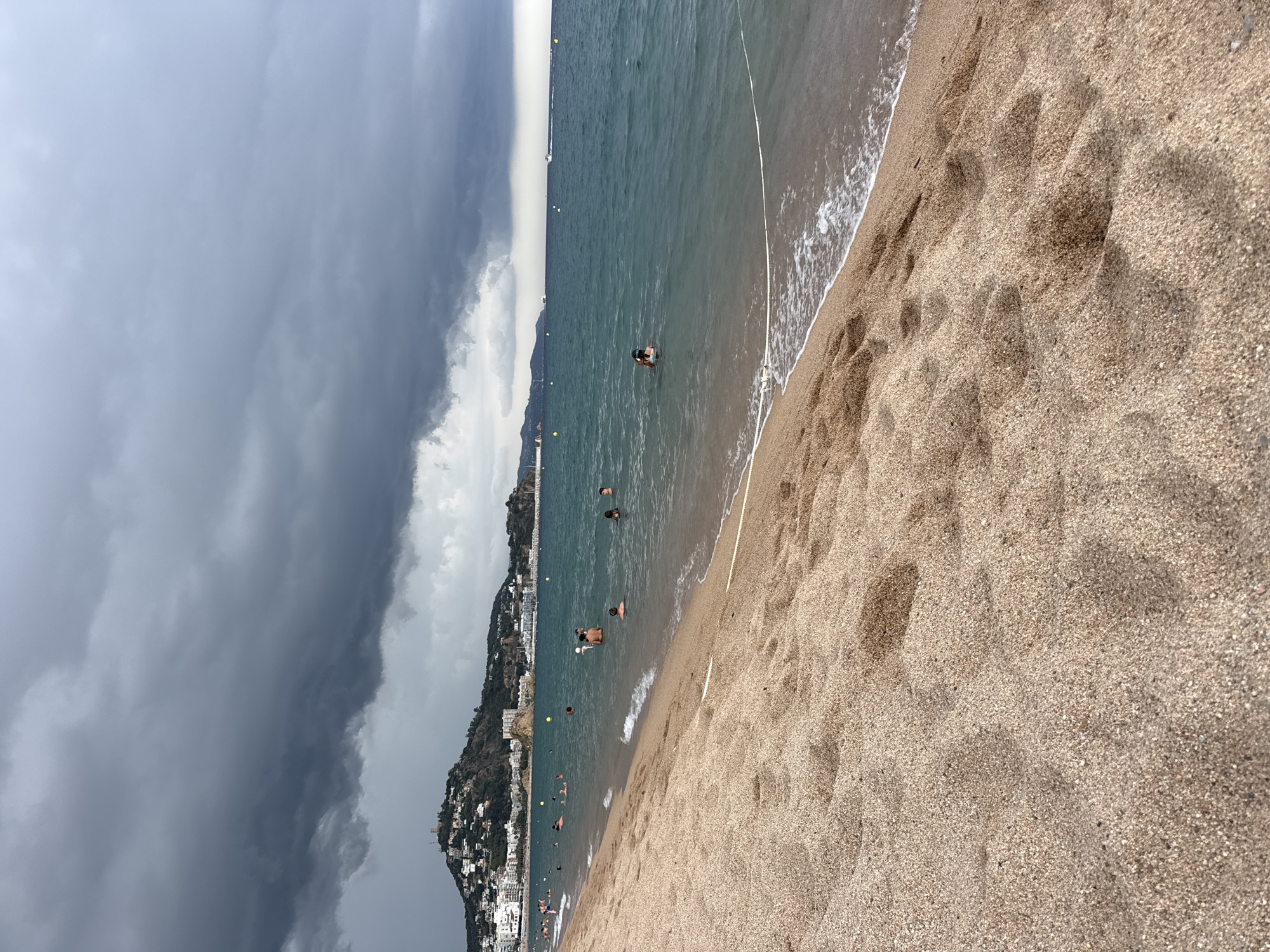 A wide-angle, low-angle shot captures a sandy beach in Blanes, Spain, on a cloudy day. The foreground is dominated by textured sand, showing subtle indentations and footprints. The water's edge is marked by gentle waves lapping onto the shore, with a white rope delineating a swimming area. In the mid-ground, several people are in the ocean, some swimming, others standing in the shallow water. The background reveals a distant town built on a hillside, topped by a prominent building that appears to be a castle or fortress. The sky is filled with dark, dramatic storm clouds, suggesting an approaching weather event. The overall scene is one of a seaside resort facing a change in weather.