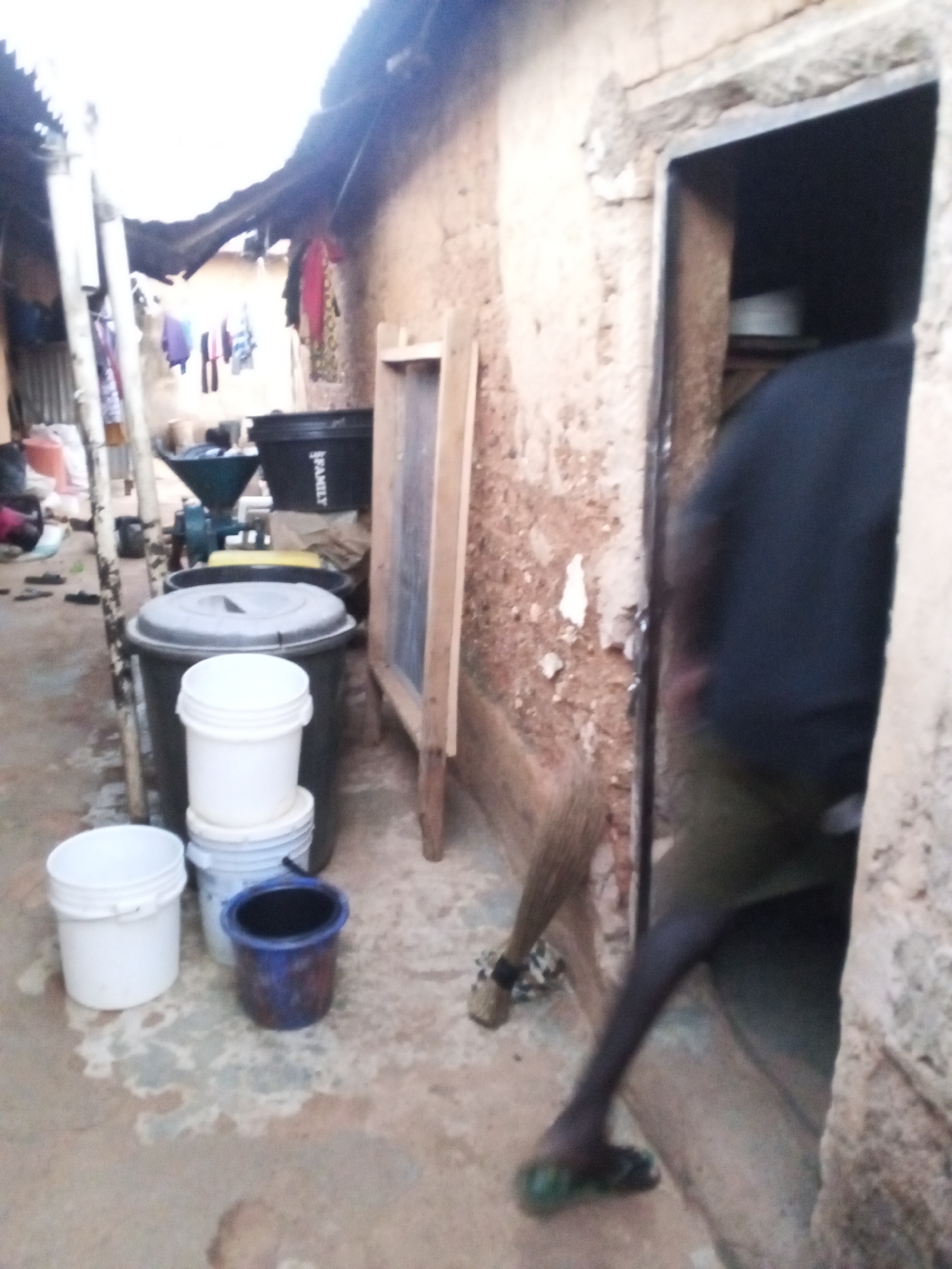 The image depicts a cluttered outdoor space in Jos, Nigeria. The walls are made of brown clay. A person is partially visible in a doorway. Multiple plastic buckets of varying sizes and colors sit on the ground near a large black drum. A grain mill is on the ground and various items of clothing hang in the background. The ground appears wet.