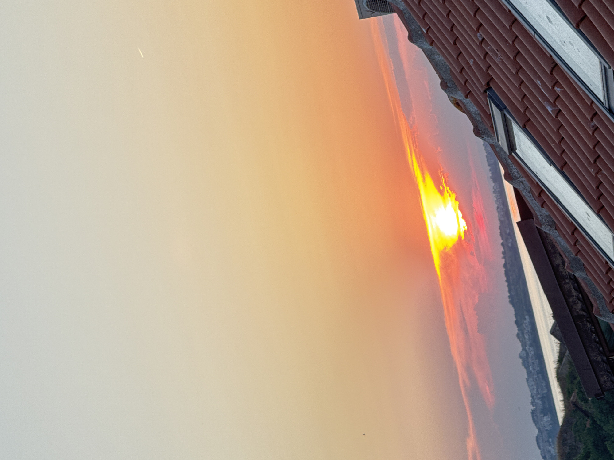 The image depicts a sunset over the city of Sozopol, Bulgaria. The sky is ablaze with warm hues of orange, yellow, and pink, with the sun partially obscured by clouds. In the foreground, a terracotta tiled roof with skylights is visible, suggesting a residential building. Beyond the roof, a body of water, likely the Black Sea, stretches towards the horizon. The distant coastline of Sozopol is dotted with white buildings, partially illuminated by the setting sun. A small, dark bird is visible in the upper left quadrant of the sky, and a faint contrail from an aircraft is present in the upper right. An outdoor air conditioning unit is mounted on the roof in the upper right corner. The overall scene captures the serene beauty of a coastal sunset.