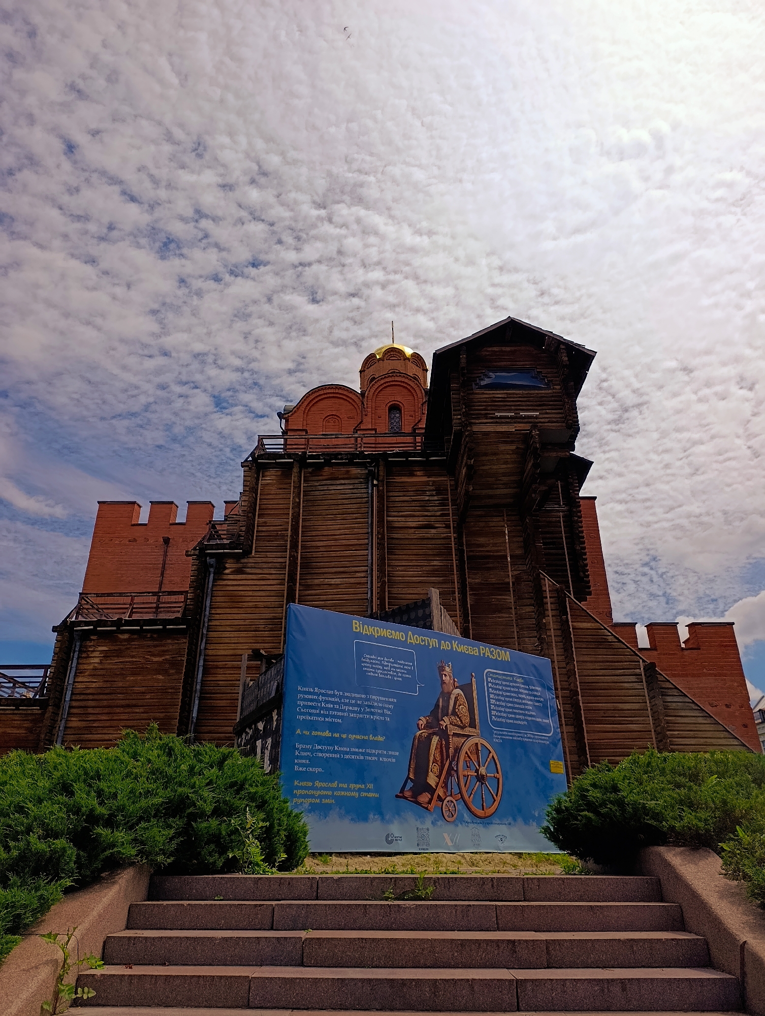 The image shows the Golden Gate of Kyiv, Ukraine, a historic landmark. The gate is made of wood and brick, with a golden dome visible at the top. In front of the gate is a large blue banner with text and an image of a person in a wheelchair, depicting a message about accessibility. The banner is placed on a pathway leading up to the gate. The sky is partly cloudy, with a few birds visible in the distance. Lush green bushes flank the steps leading to the gate. The perspective is low-angle, looking up at the gate and the sky.