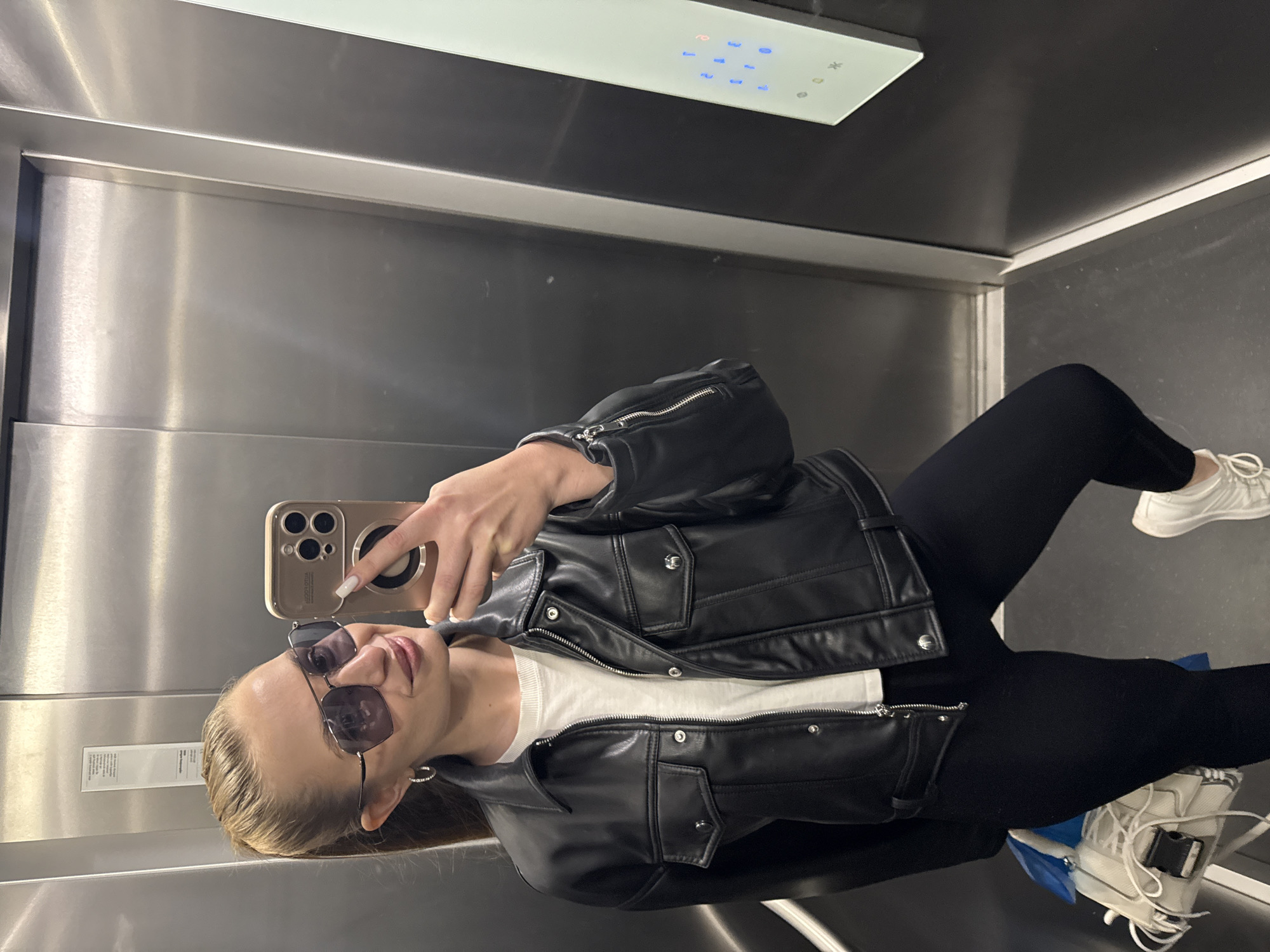 A woman takes a selfie in an elevator. She wears sunglasses, a black leather jacket, white shirt, black leggings, and white sneakers. The elevator is a modern design with stainless steel walls and a white panel with lit buttons. The location is an elevator in a building in Budva, Montenegro.