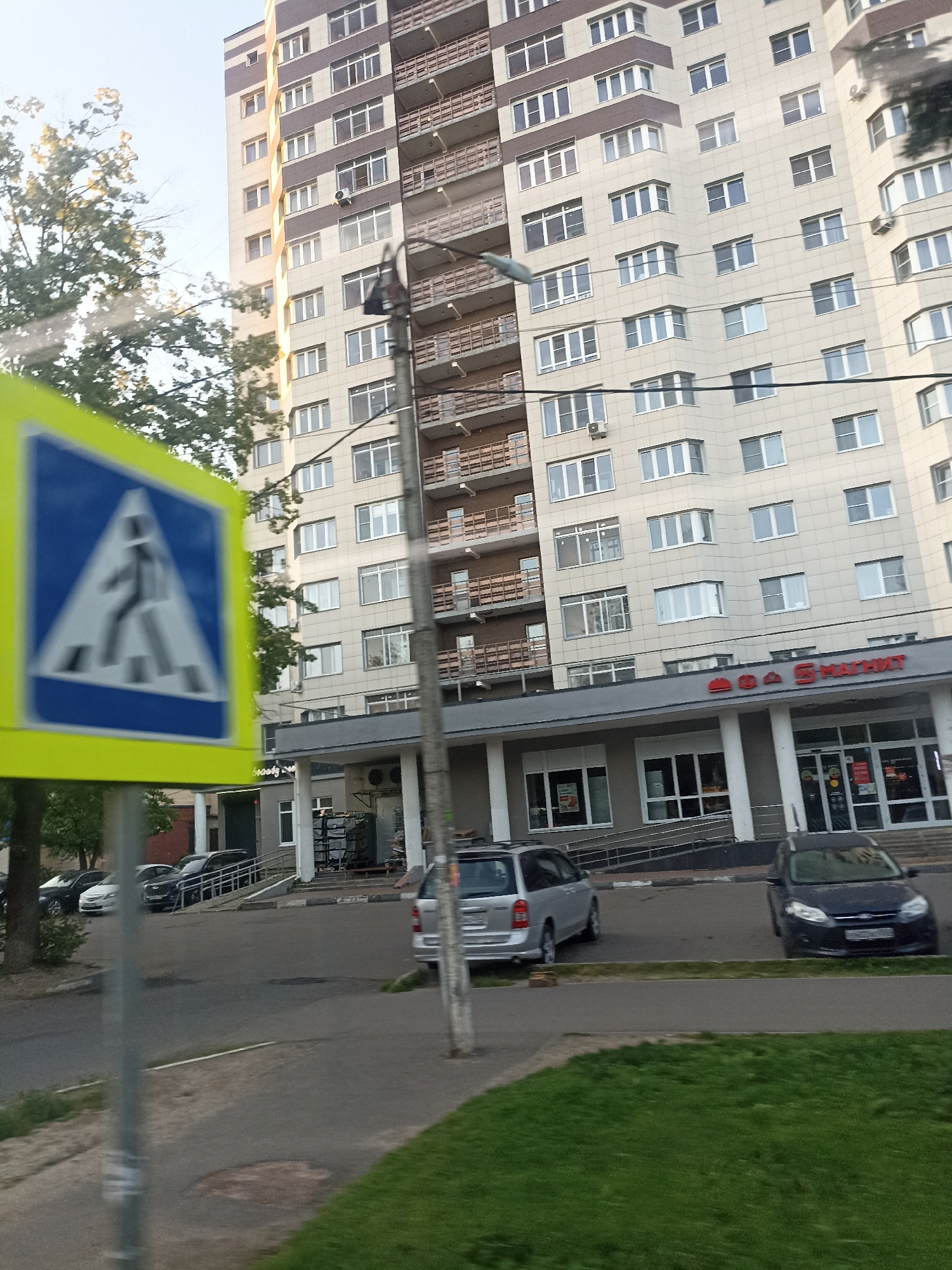 The image depicts a street scene in Chekhov, Russia, with a focus on a modern apartment building and surrounding urban elements. The apartment building is tall and multi-story, with a beige facade and brown accents. It features numerous windows and balconies. In the foreground, a pedestrian crossing sign with a blue triangular sign and a yellow border is prominently displayed, indicating a crosswalk. Several cars are parked on the street, including a dark sedan, a silver minivan, and other vehicles. A supermarket with a red and white sign is visible at the base of the apartment building. The environment includes trees and a grassy area, suggesting a developed residential or commercial area. The lighting suggests it is daytime, possibly late afternoon or early morning given the soft light.