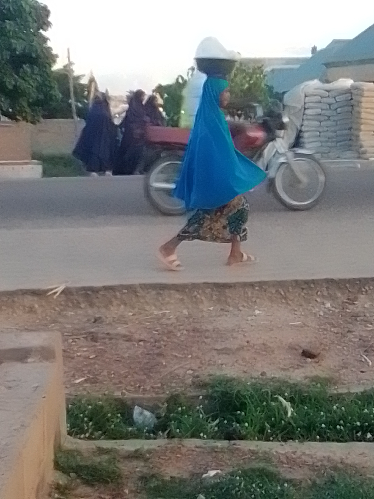 In the city of Gombe, Nigeria, a woman walks along a paved road, carrying a large, white bowl of goods on her head. She is wearing a vibrant blue headscarf and a patterned skirt. Behind her, a motorcycle is visible, loaded with sacks of what appears to be cement. Several other figures, also in dark clothing and headscarves, are present in the background. The scene takes place outdoors, with trees and buildings lining the road. The ground in the foreground is covered in gravel and sparse vegetation.