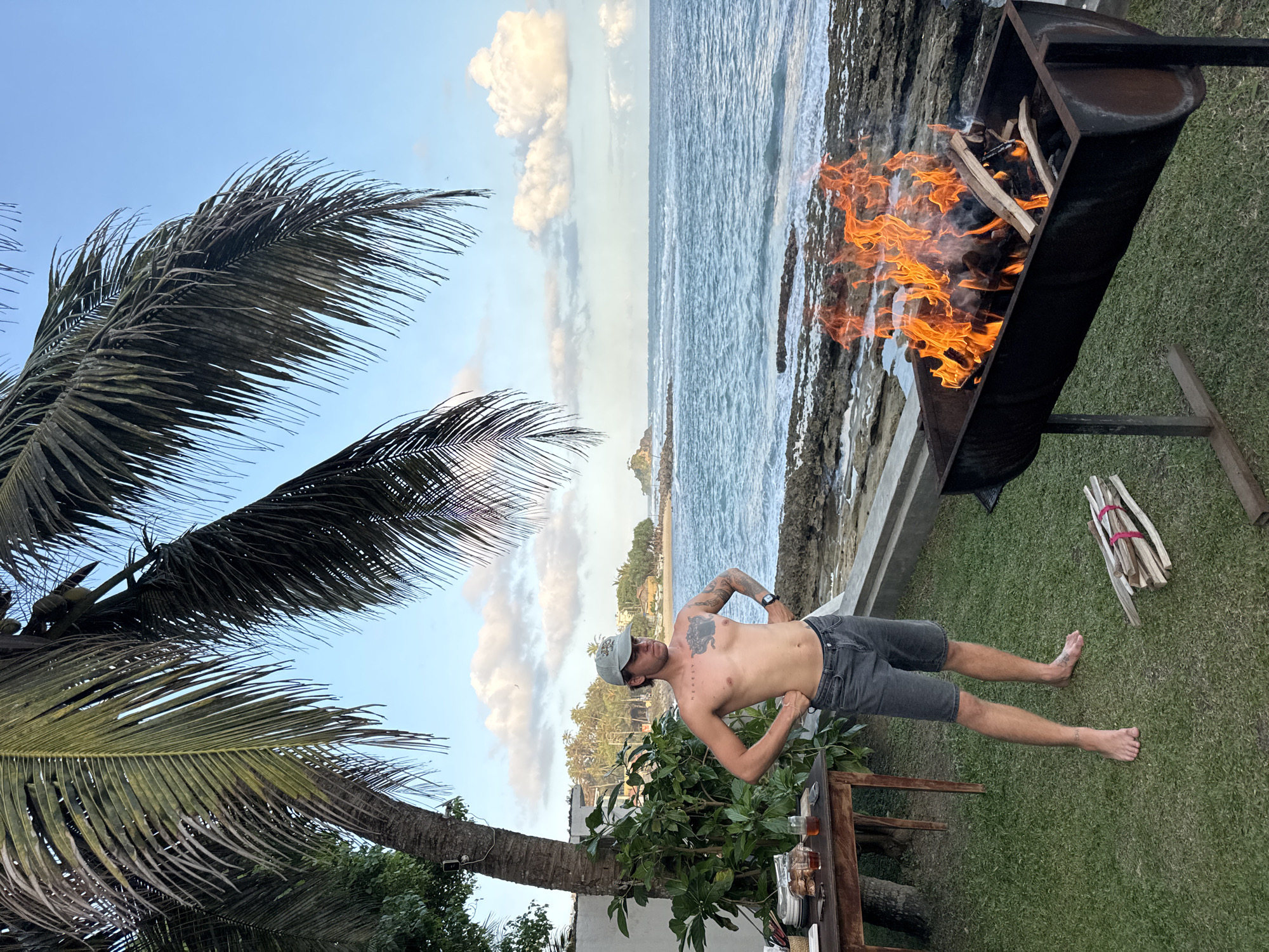 A man stands near a barbecue pit on a grassy lawn overlooking the ocean. The man is shirtless, wearing grey shorts and a cap. His hands are on his hips and he has tattoos on his chest and arms. In the foreground, a barbecue pit contains a roaring fire. To the left, a wooden table is set with plates and drinks. The background shows the ocean with waves crashing against rocks, and a distant island with buildings and palm trees. The sky is blue with scattered clouds. The scene appears to be set in Goviyapana, Sri Lanka.