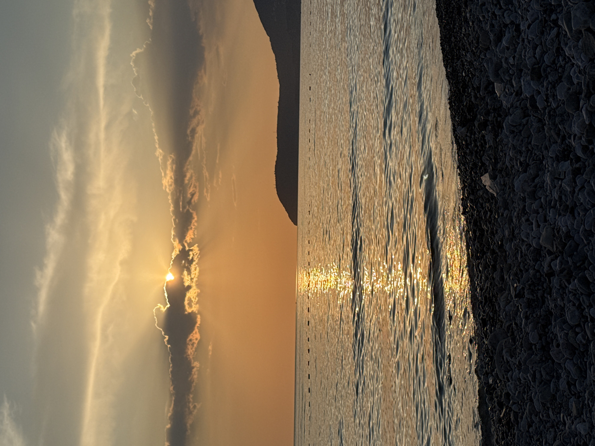 The image captures a beach scene in Bar, Montenegro at sunset. The sun is partially obscured by clouds, casting golden rays across the sky. The light reflects on the water, creating shimmering patterns. A dark, rocky coastline is visible on the right side of the image, while a silhouette of a land mass can be seen in the distance. The scene is devoid of people.