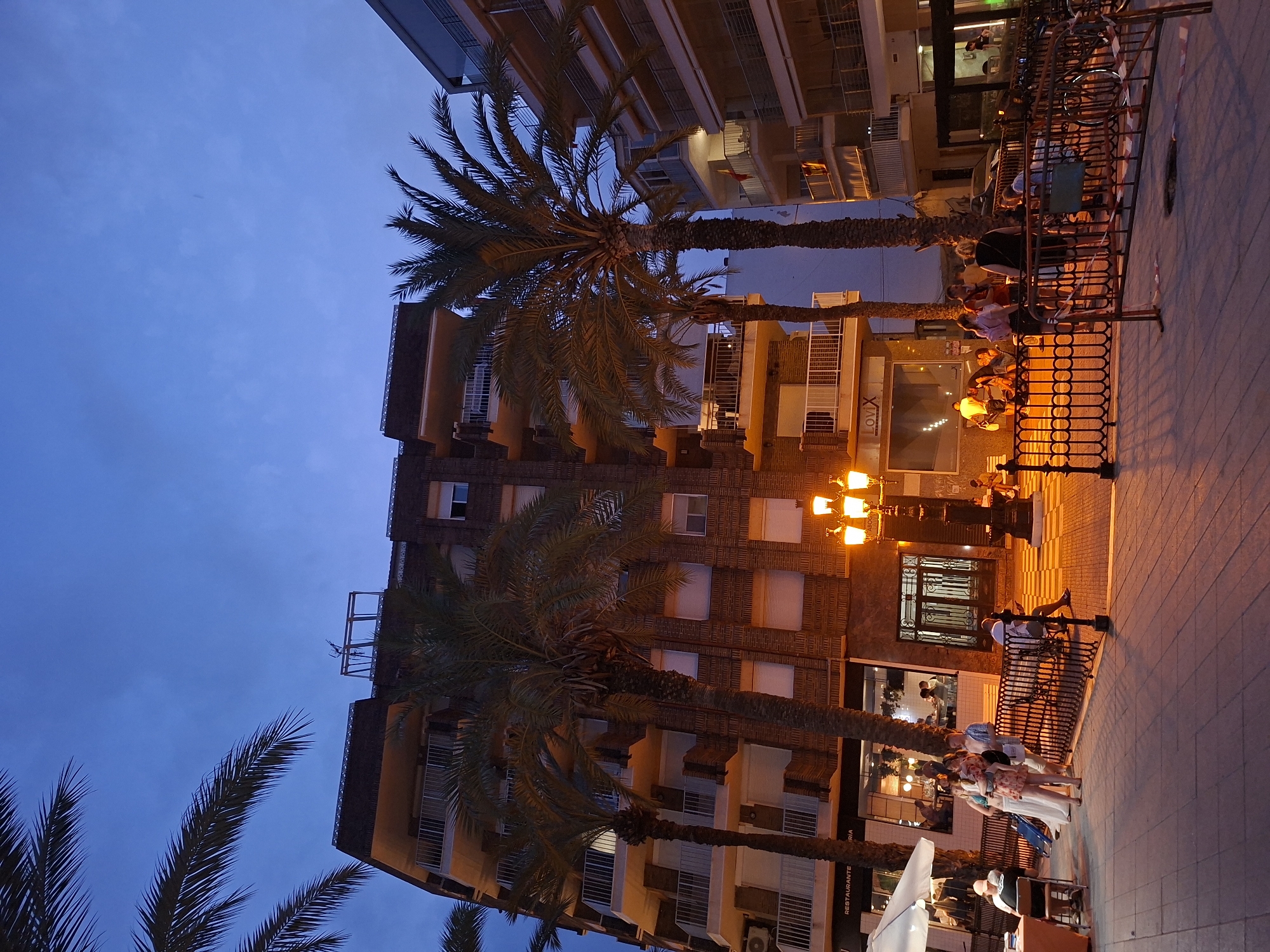 The image depicts a street scene in Torrevieja, Spain, at dusk. In the foreground, a paved walkway is illuminated by streetlights, with people seated on benches and standing in small groups. A decorative black metal fence borders the walkway, with some caution tape visible. Palm trees are scattered throughout the scene. Behind the walkway, a multi-story building constructed of brick with balconies and windows is visible. To the right, another multi-story building with balconies and white railings is present. Businesses with illuminated windows are visible on the ground floor of the brick building. The sky is a deep blue, indicating the transition from day to night.