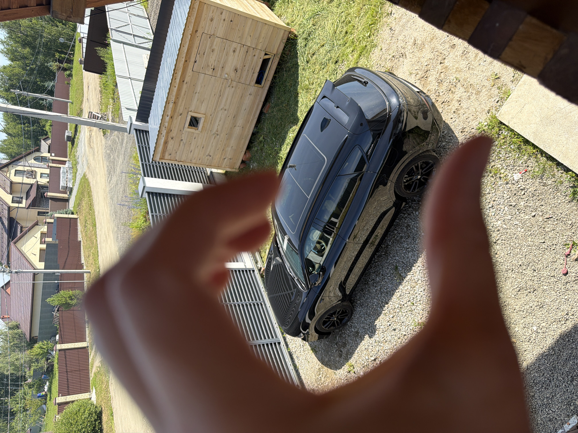 In Malinovka, Russia, a black car is parked on a gravel driveway outside a residential property. The car, identified by the "VOYAH" badge, is angled to the left, with its sunroof open. Adjacent to the car is a small, wooden structure with a corrugated metal roof, resting on a grassy area. The scene is framed by a human hand, blurred and in the foreground, appearing to form a heart shape. Beyond the property, residential buildings and trees are visible, suggesting a suburban or rural setting. The sky is clear and the sunlight indicates a daytime scene.