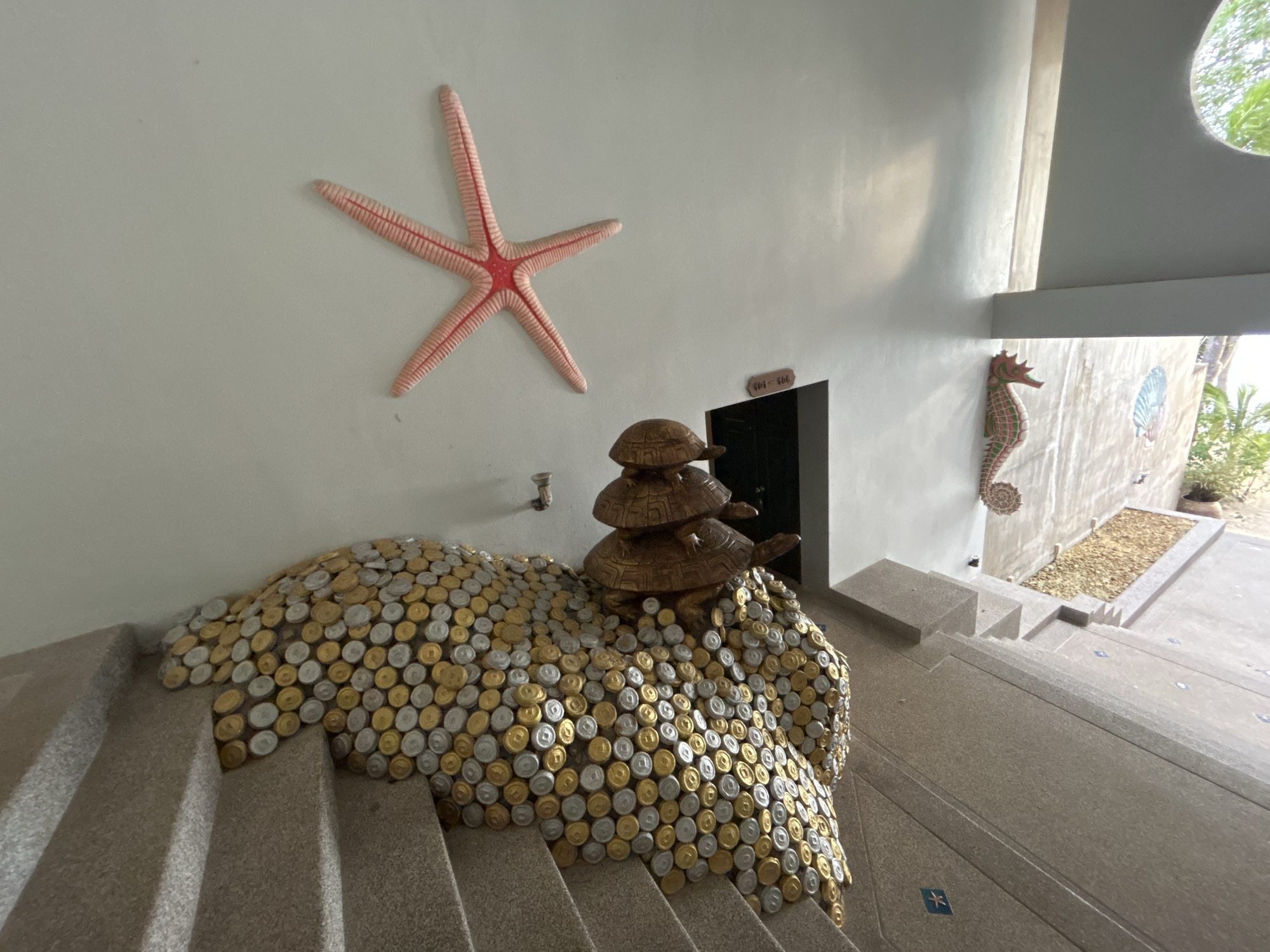 The image depicts an indoor stairway in ตำบลเกาะพะงัน, Thailand, decorated with nautical-themed art. On the left wall, a large red and white starfish is mounted, positioned above a sculpture composed of a mound of gold and silver bottle caps cascading onto the stairs. A stack of three wooden turtles rests atop the bottle cap mound. In the background, a door labeled "404-406" is visible, and further down the stairs, a seahorse is mounted on the wall. The stairs themselves are light grey, and the walls are a light, neutral color. The lighting is a mix of natural light from an unseen source and artificial light within the building. The scene appears to be an entrance or lobby area.