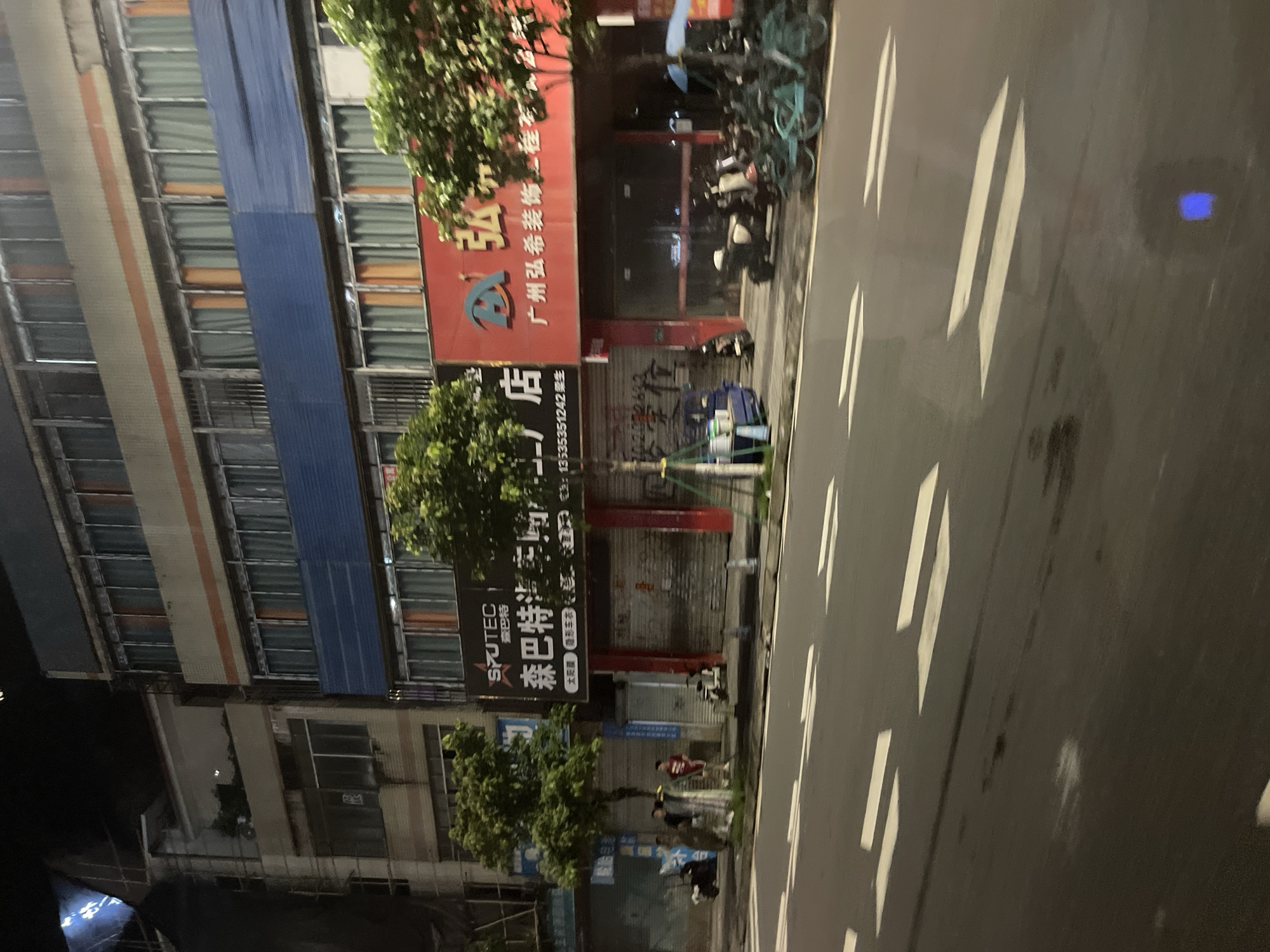 The image captures a street scene at night in Panyu District, China, focusing on the facades of several buildings lining the road. The buildings appear to be multi-story commercial structures. On the ground floor, various shops are visible with closed metal roll-up doors. Above, windows with closed curtains, some orange and some greenish-blue, are prominent across multiple levels of the buildings. A red building with white and blue signage is particularly noticeable, featuring Chinese characters and a logo. Trees with green foliage are planted along the sidewalk in front of the shops. Parked on the sidewalk and street are several motorcycles and bicycles, including some teal-colored bikes. A blue cart with white buckets sits on the sidewalk. A few individuals, appearing to be young men, are standing or walking on the sidewalk. The street in the foreground is wet and reflects the surrounding lights. White directional arrows are painted on the road surface. The overall atmosphere is that of a busy commercial street at night, with businesses closed.