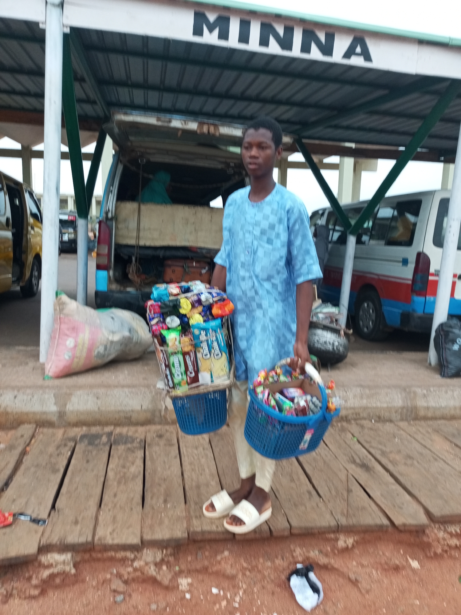 A young boy stands in front of a covered stall with the word "MINNA" written in large black letters above. He wears a light blue traditional outfit and white sandals. In his hands, he holds two blue baskets filled with various packaged snacks and sweets. A large pink and gray sack lies on the ground to his left. Behind him, a van with red and blue stripes is parked under the shelter. Other vehicles are visible in the background. The ground is a mix of wooden planks and reddish-brown soil. The scene appears to be an outdoor market or transportation hub in Gombe, Nigeria.