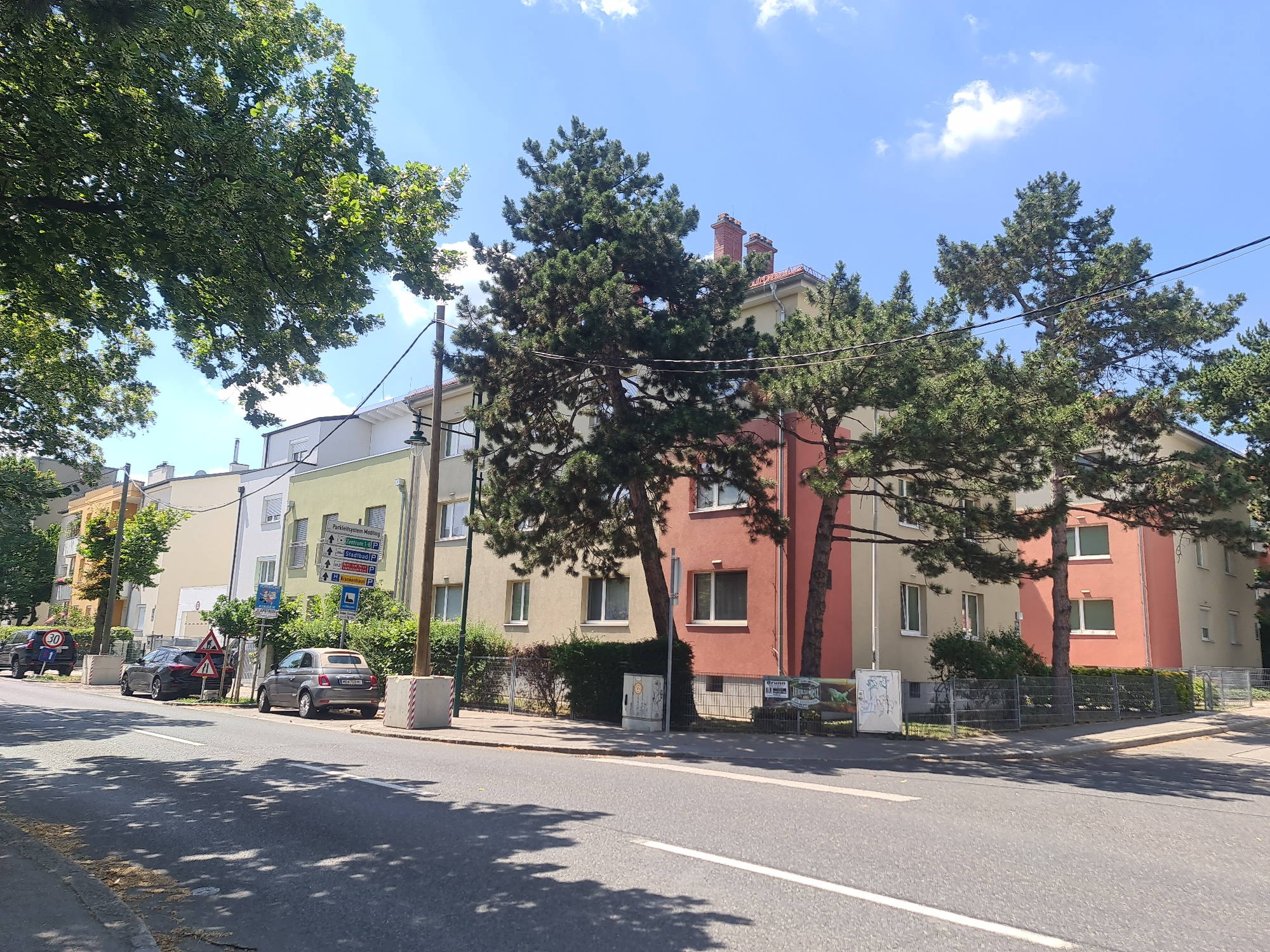 This is a street-level, daytime, eye-level, medium shot of a residential street in Mödling, Austria. The scene depicts a row of multi-story apartment buildings with pastel-colored facades, some with red brick accents. Large evergreen trees with dense foliage are situated in front of and between the buildings, casting shadows on the street and sidewalk. A paved road runs diagonally across the foreground, with a white line marking the lane. Parked cars, including a gray Fiat 500 and a dark sedan, are visible along the curb. Several street signs are present, including one with multiple directional arrows and speed limit signs. The sky is blue with scattered white clouds. The overall impression is a typical urban residential setting on a sunny day.