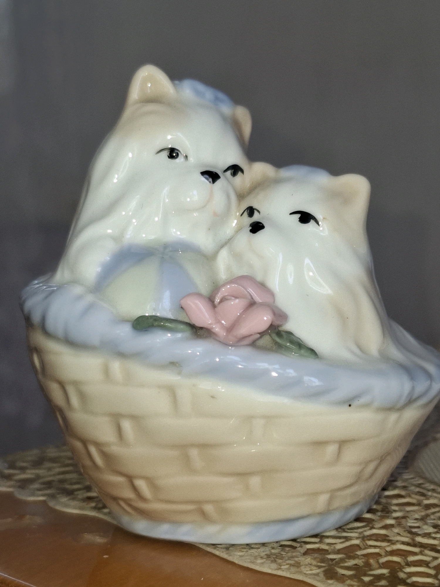 A ceramic figurine of two white cats nestled in a basket. The cats have blue accents on their ears and the basket has a light tan woven texture with a light blue trim. A small pink rose and a green leaf are in front of the cats. The figurine is placed on a brown surface with a patterned mat. The background is a dark gray. The scene is indoors and there is no information about the location in Almaty, Kazakhstan.