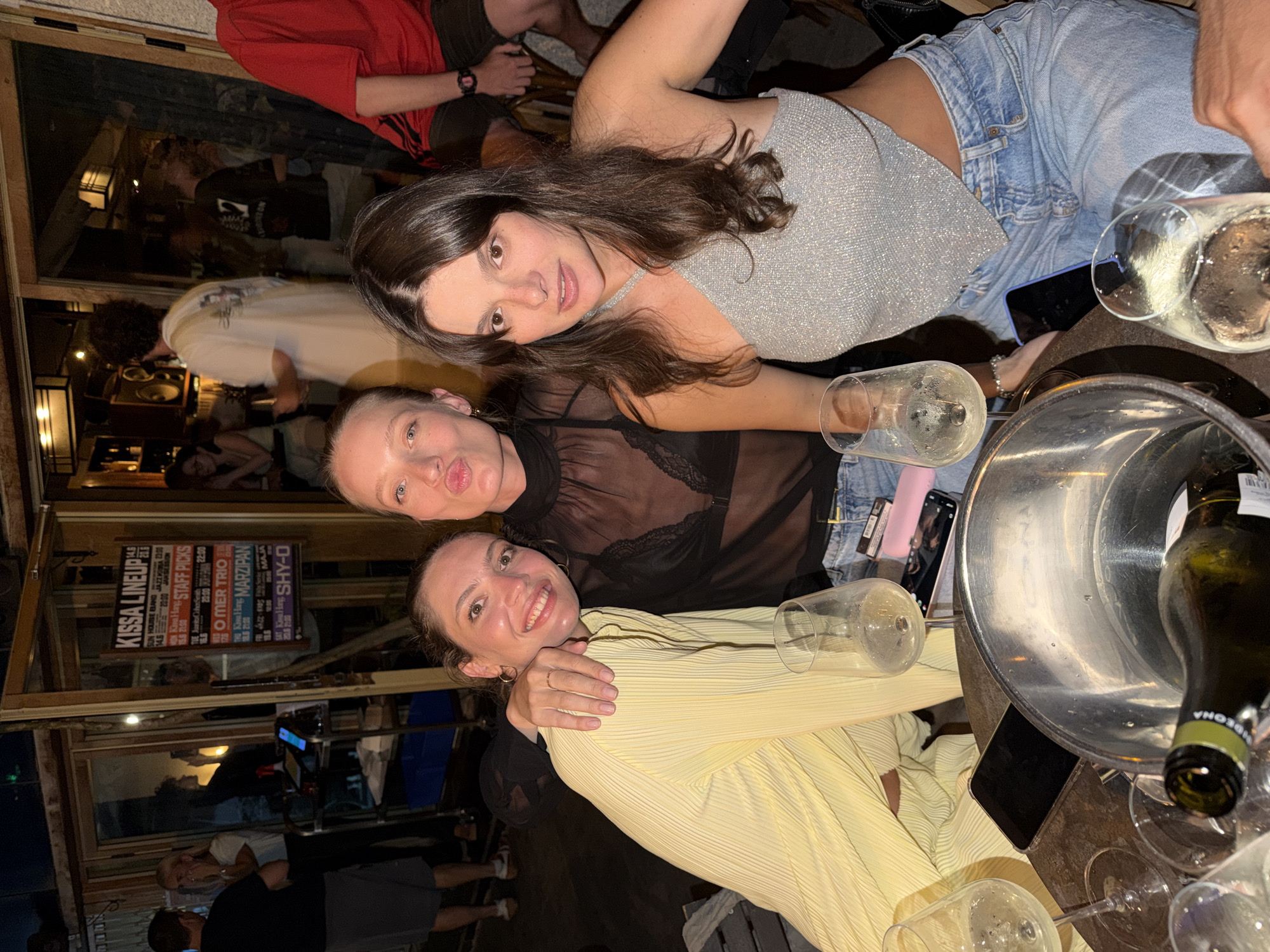 Three young women are posing for a photograph at an outdoor bar or restaurant in Tel Aviv, Israel. The woman on the left is wearing a yellow pleated dress and has her arm around the woman in the middle. The woman in the middle is wearing a sheer black top and jeans, and she is puckering her lips for the camera. The woman on the right is wearing a silver sparkly halter top and jeans and is looking at the camera. They are sitting at a table with drinks, a bottle of wine in an ice bucket, and mobile phones. In the background, other patrons and a sign for a "Kissa Lineup" can be seen.