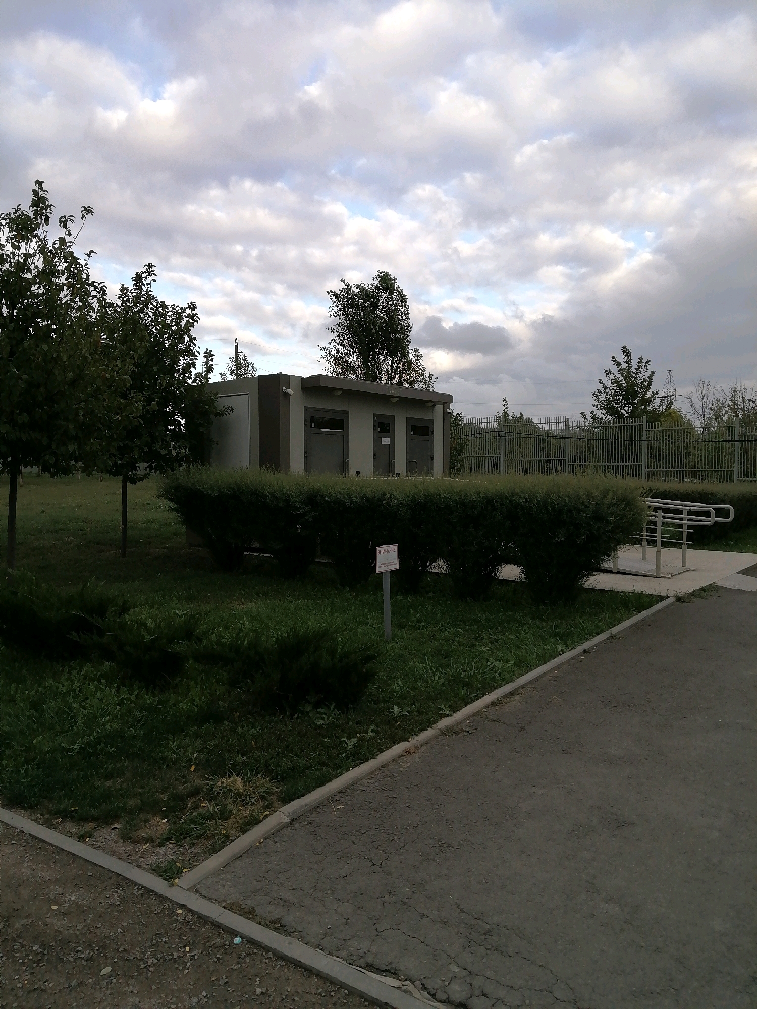 The image shows an outdoor scene with a modern building in the background, a manicured hedge in the foreground, and trees on the left. The sky is overcast with clouds. To the right of the building, there is a metal railing and stairs leading up to a paved area. A small sign is visible in front of the hedge. The location is identified as being in the city of Rostov-on-Don, Russia. The scene appears to be a public space or park.