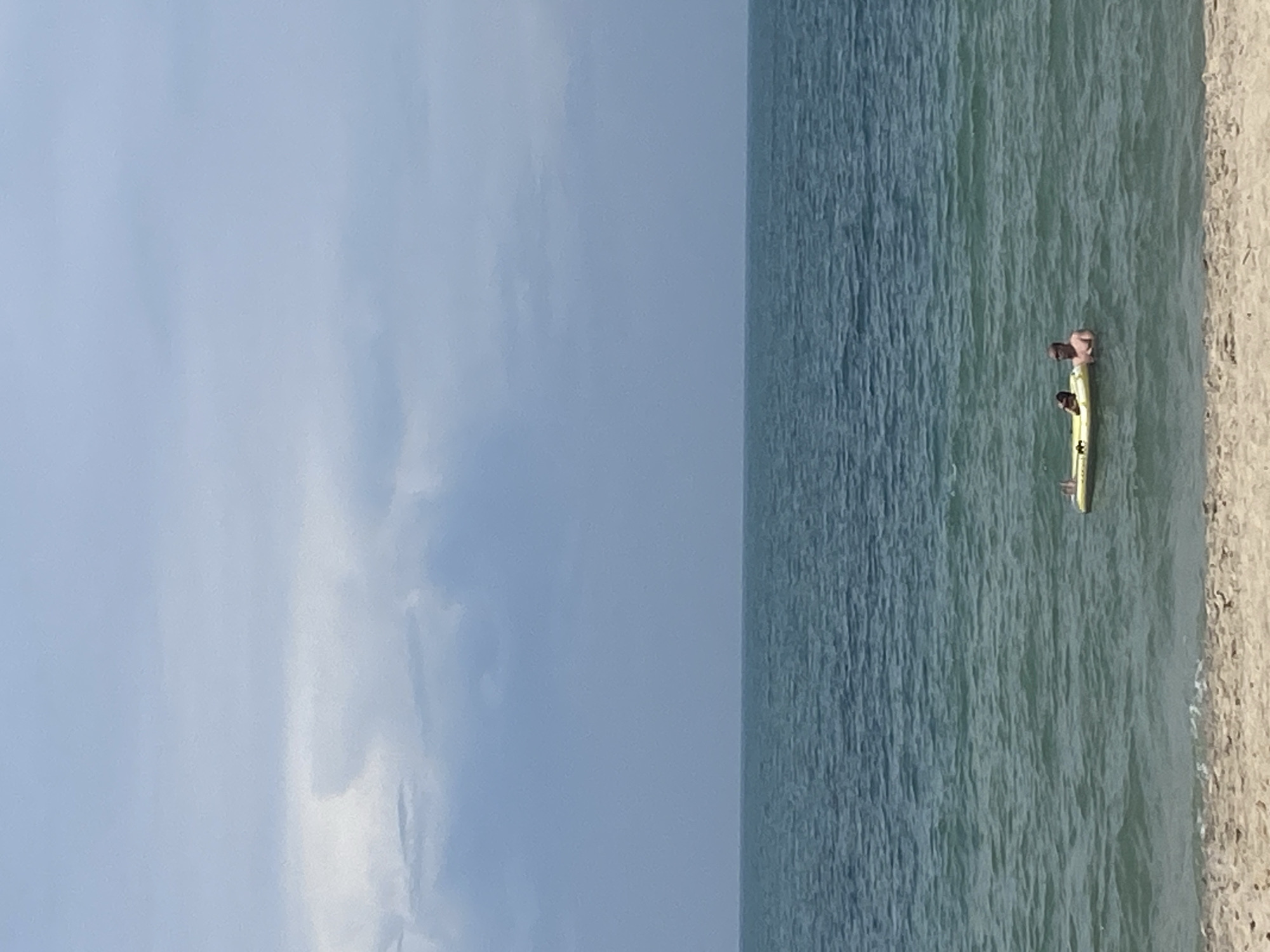 Two individuals are seen on a yellow inflatable raft in the ocean. The raft is positioned in the middle of the frame, with the water extending from the foreground to the horizon. The sky above is overcast, with clouds covering the upper portion of the image. The foreground shows a sandy beach. The context of Phu Thuy Ward, Vietnam is not discernible from the image itself.