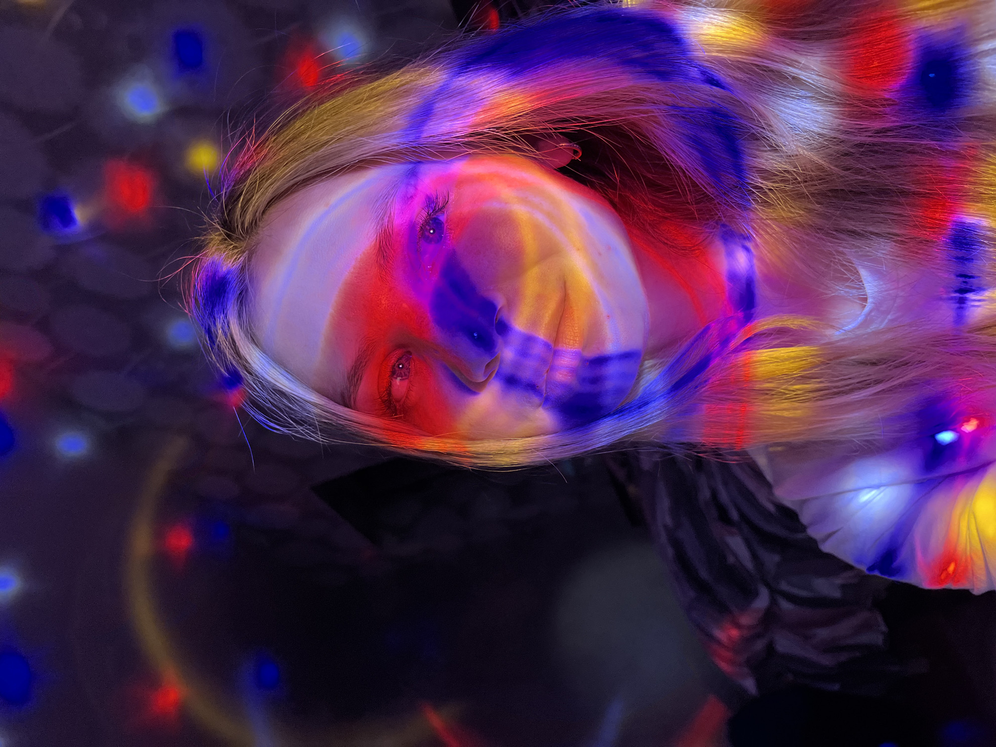 A woman with fair skin and blonde hair is depicted in a close-up shot. Her face is illuminated by colorful lights, predominantly red, blue, and yellow, creating a surreal effect. The lights appear to be projected onto her face and hair, giving them vibrant hues. Her eyes are visible, appearing red due to the lighting. The background is dark and blurred, with scattered light spots suggesting an urban setting at night. Based on the query, the location is identified as the city of Chaikovsky, Russia. The scene appears to be artistic or staged, possibly involving a light display or a photographic effect.
