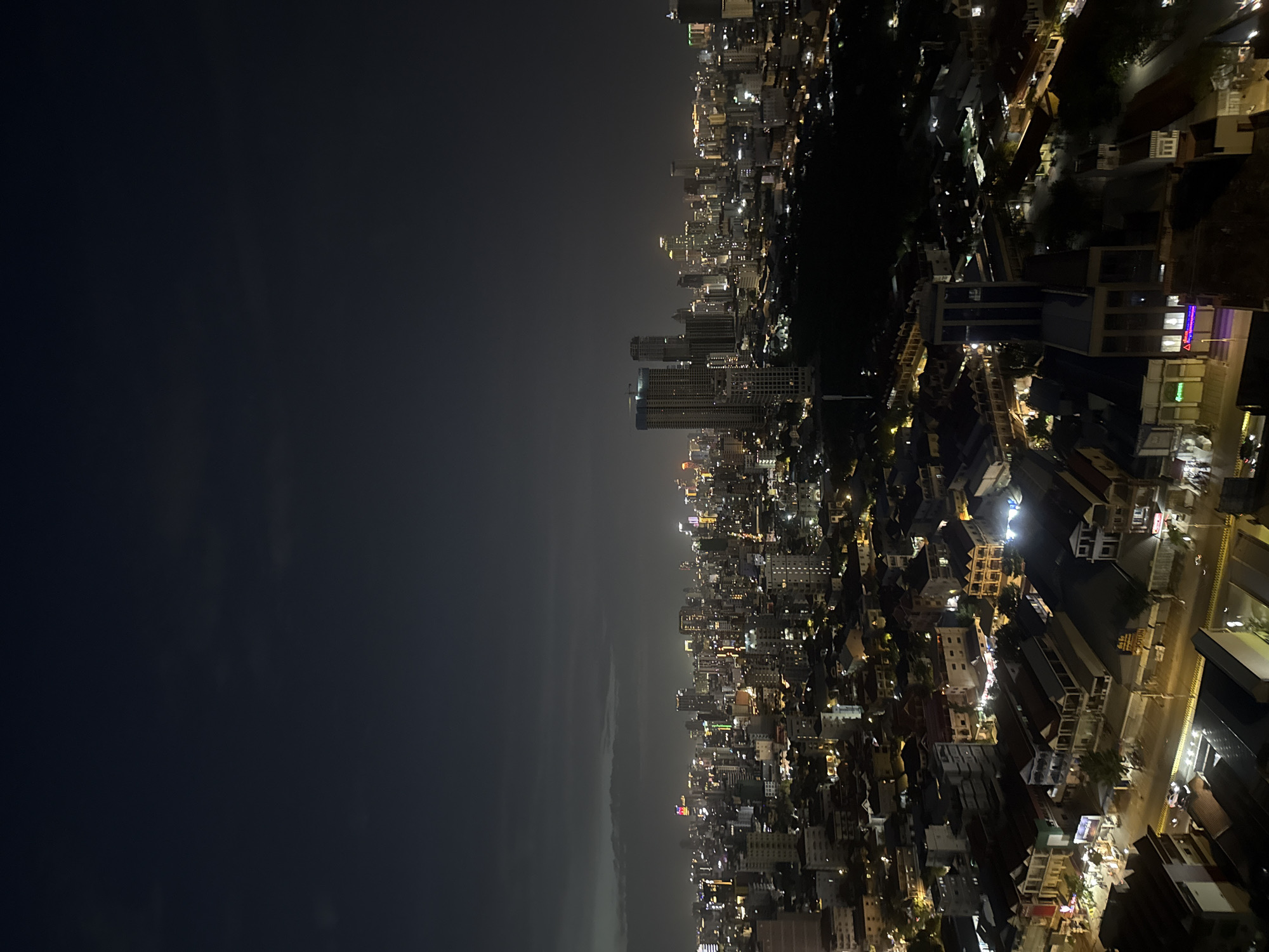 This is a nighttime, high-angle, wide shot of a cityscape, likely taken from a tall building in Khan Mean Chey, Cambodia. The sky is dark and overcast, with faint clouds visible towards the horizon. The city below is illuminated by a dense network of artificial lights, creating a vibrant urban scene. Tall skyscrapers with illuminated windows dominate the background, interspersed with numerous lower-rise buildings, many with brightly lit facades. Streets are visible, with lines of lights suggesting traffic and streetlights. The foreground shows more detailed views of buildings, including residential structures with visible rooftops and some commercial establishments with illuminated signs. The overall impression is of a bustling, densely populated city at night.