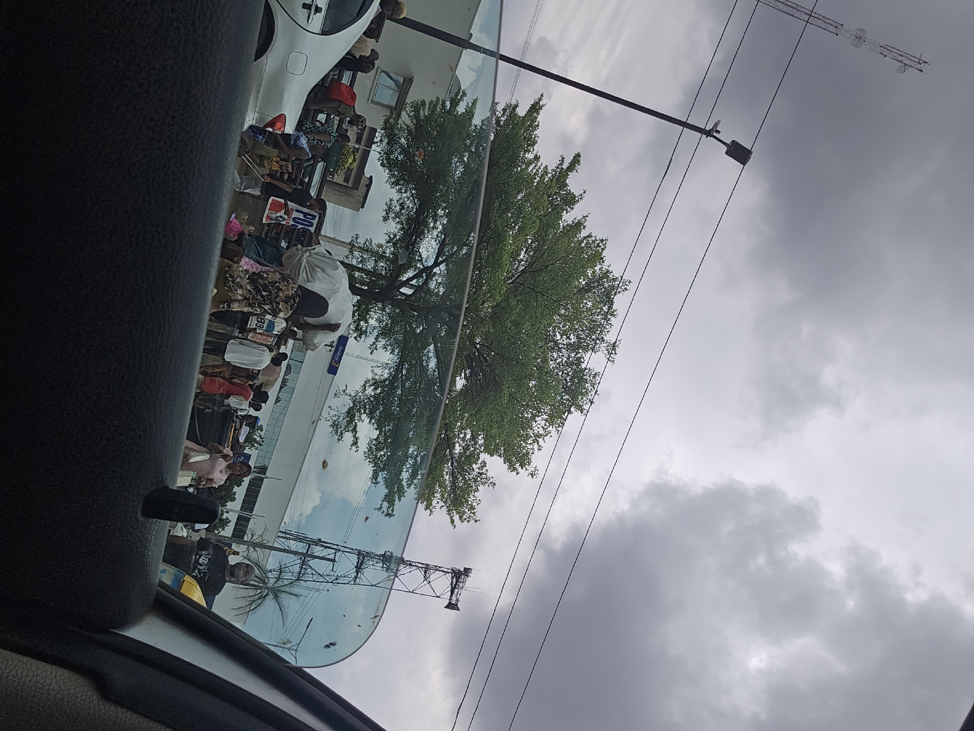 The view from inside a car in Benin City, Nigeria, shows a reflection in the windshield of a busy street. People stand along the sidewalk with goods displayed for sale; a white car is parked along the road. The sky is overcast. Overhead electrical wires run across the image. In the reflection, a communication tower can be seen. A large tree stands near the curb.