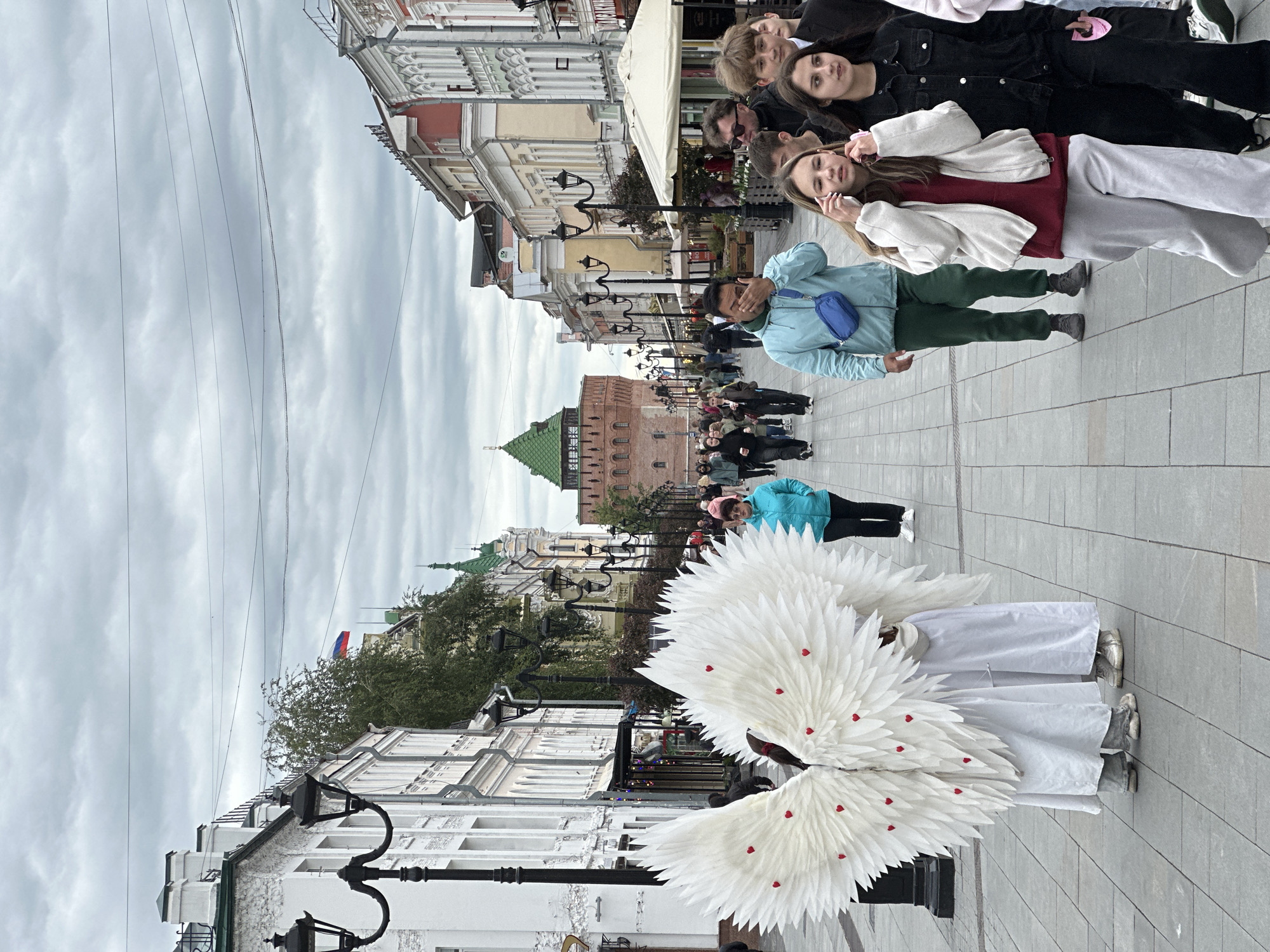In Nizhny Novgorod, Russia, the image depicts a pedestrianized street with historic buildings on either side under an overcast sky. Dominating the scene are two individuals in white costumes with large white feather wings adorned with red heart-shaped accents. They are positioned on the paved street, which appears to be a popular promenade.
Further down the street, a group of people are walking, with some individuals posing for photos. A man in a light blue jacket is positioned between the camera and the crowd, partially obscuring his face. In the background, a prominent brick tower with a green roof is visible, likely part of the Nizhny Novgorod Kremlin. Several lampposts line the street, and overhead wires stretch across the sky. The Russian flag is discernible among the buildings on the left side of the image.