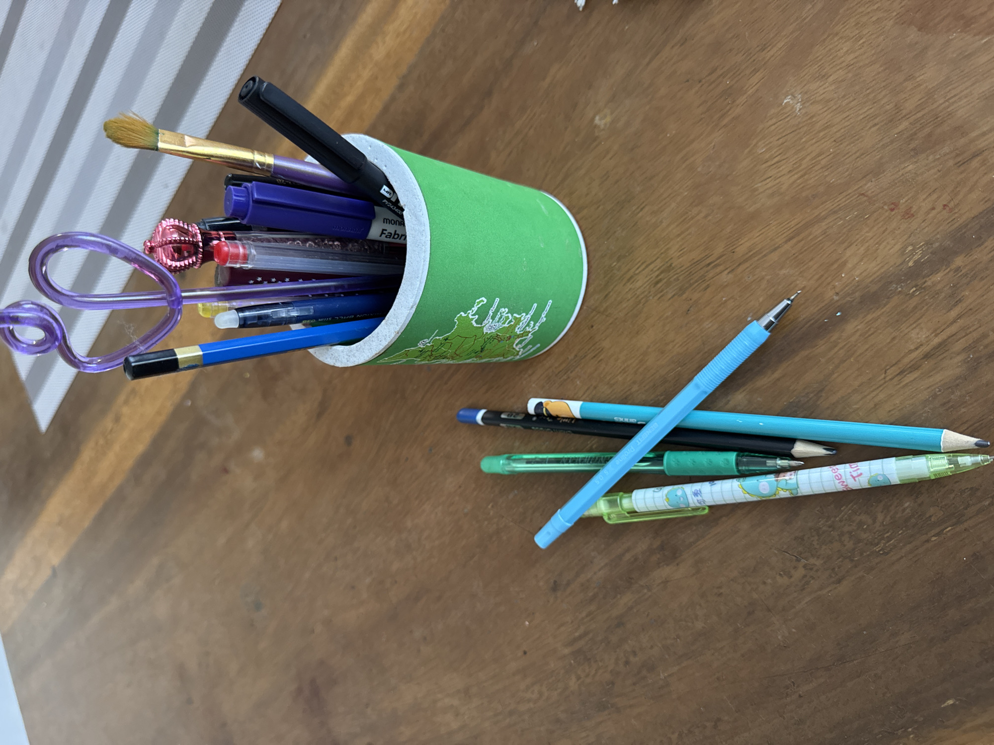 The image shows a collection of writing and art supplies on a wooden table. A green, cylindrical container with a map design is filled with pens, pencils, a paintbrush, and a whimsical, spiraled drinking straw. To the right of the container, on the table, are several additional pencils and pens scattered. The table surface is a medium brown color with visible wood grain. A striped surface is visible in the upper left corner, possibly a placemat or table runner. The scene appears to be indoors. There is no evidence in the image to indicate the location is Kuta Selatan, Indonesia.