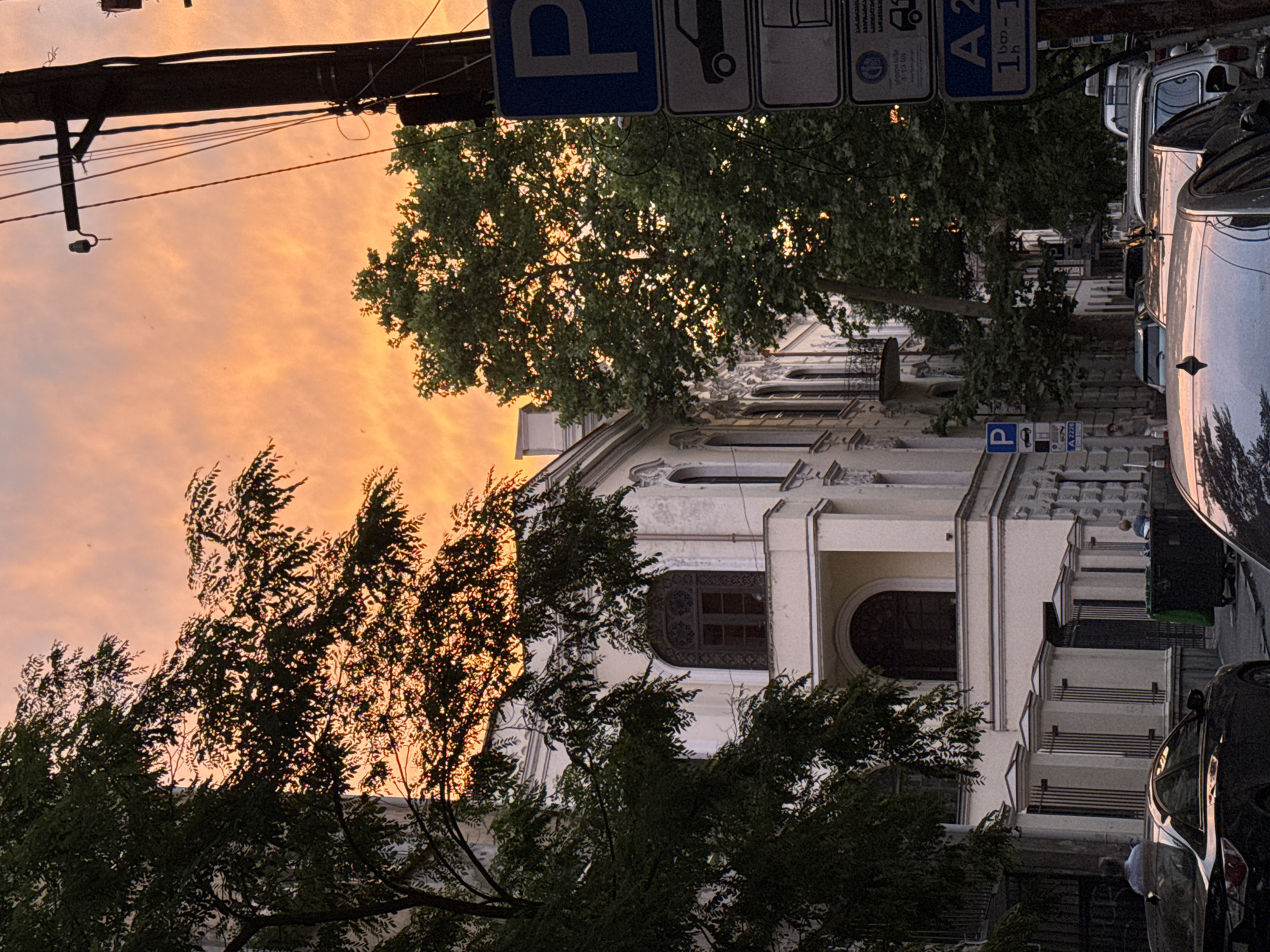 The image depicts a street scene in Tbilisi, Georgia, likely during sunset. The sky is ablaze with the warm hues of orange and pink, suggesting the golden hour. A prominent utility pole stretches across the top of the frame, adorned with wires and what appears to be a small light fixture. Below, a lush green tree with dense foliage partially obscures a light-colored, multi-story building. The building features an ornate facade with decorative elements and several windows. In the foreground and to the right, parked cars are visible, one of which is dark and shiny, reflecting the surrounding environment. Several street signs are also present, including a blue "P" sign indicating parking. One sign has text in what appears to be Georgian script. The overall atmosphere is serene, with the vibrant sunset creating a warm and inviting ambiance. The wind seems to be rustling the leaves of the trees, adding a sense of movement to the scene.