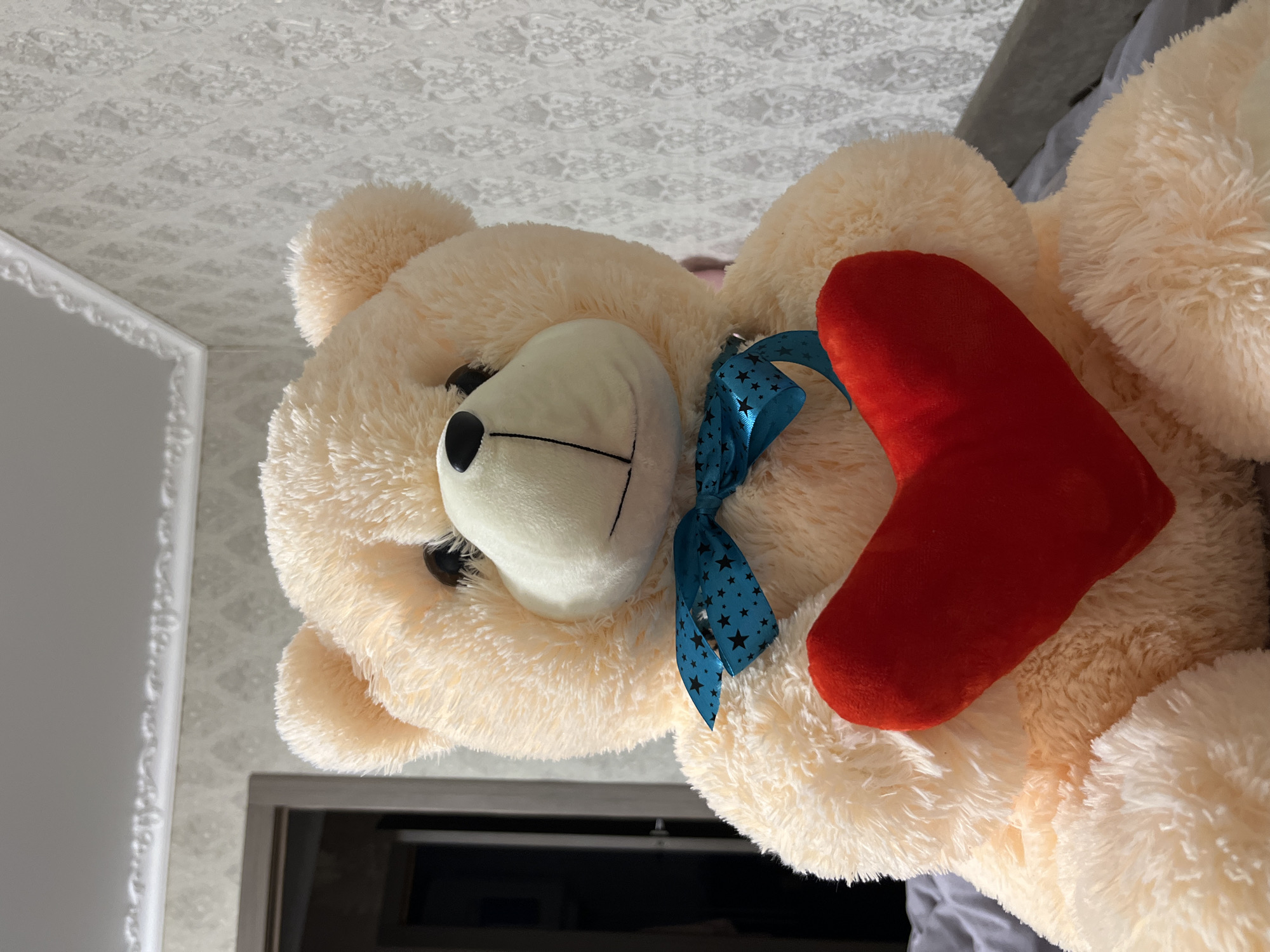 A close-up shot depicts a large, cream-colored teddy bear holding a red heart. The bear has a white muzzle, black eyes, and a blue ribbon with black stars tied around its neck. The bear's fur is fluffy. The background includes a patterned white ceiling and a glimpse of a dark-colored doorway.