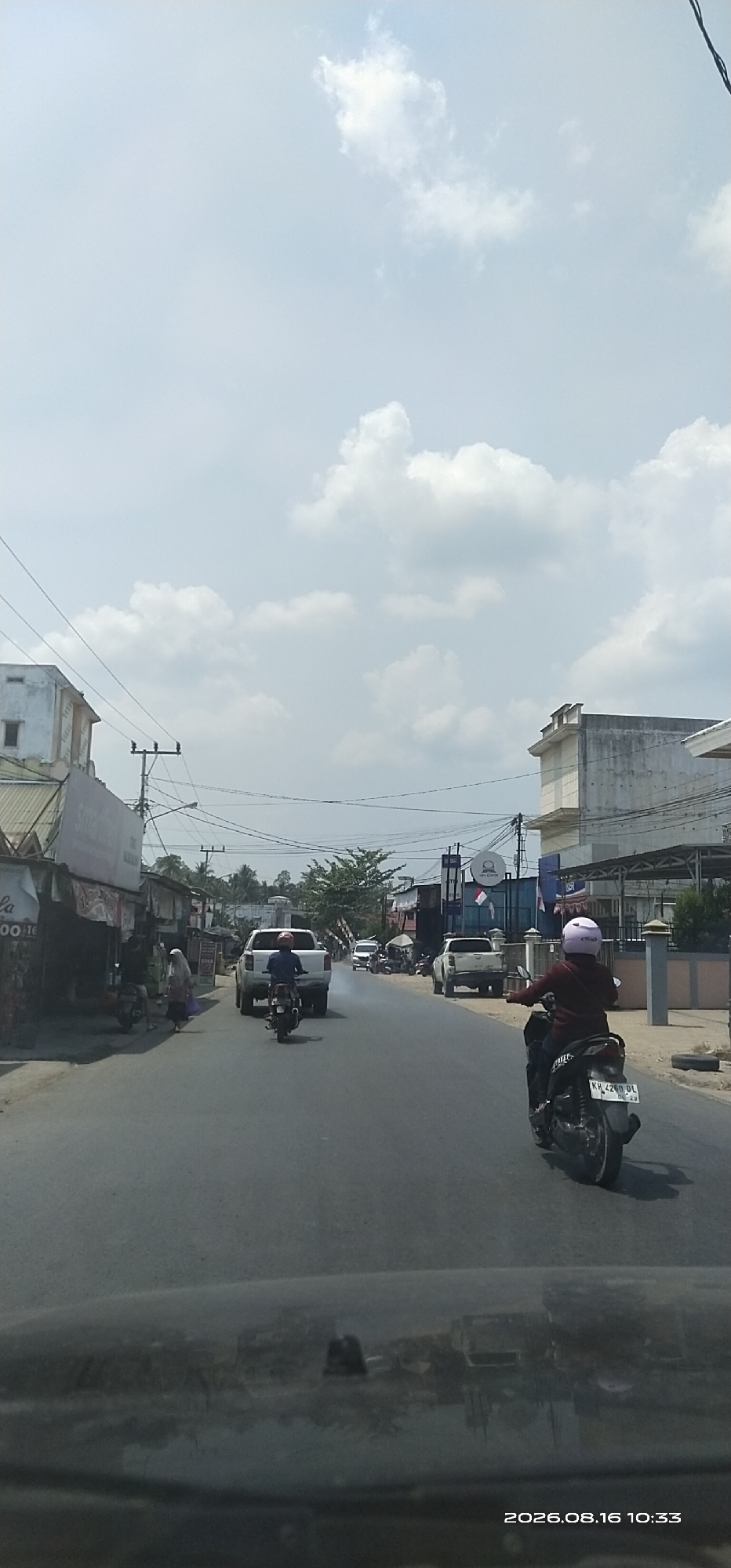 The image depicts a street scene in Ampah Kota, Indonesia. In the foreground, the dashboard of a vehicle is visible, reflecting the street and surrounding buildings. A motorbike with a rider wearing a maroon jacket and a light purple helmet is in the right lane, traveling away from the viewer. The motorbike's license plate reads "KH 4200 DL". Further down the road, a white pickup truck is in the left lane, also moving away. A second motorbike is in the left lane ahead of the pickup, also traveling away.
On the left side of the street, a row of businesses with awnings and signage lines the road. A woman in a headscarf is seen walking on the sidewalk in front of one of these businesses. On the right side of the street, more buildings and businesses are visible, with some parked motorbikes. Power lines and utility poles are present throughout the scene. The sky is partly cloudy. The timestamp "2026.08.16 10:33" is displayed in the bottom right corner.
