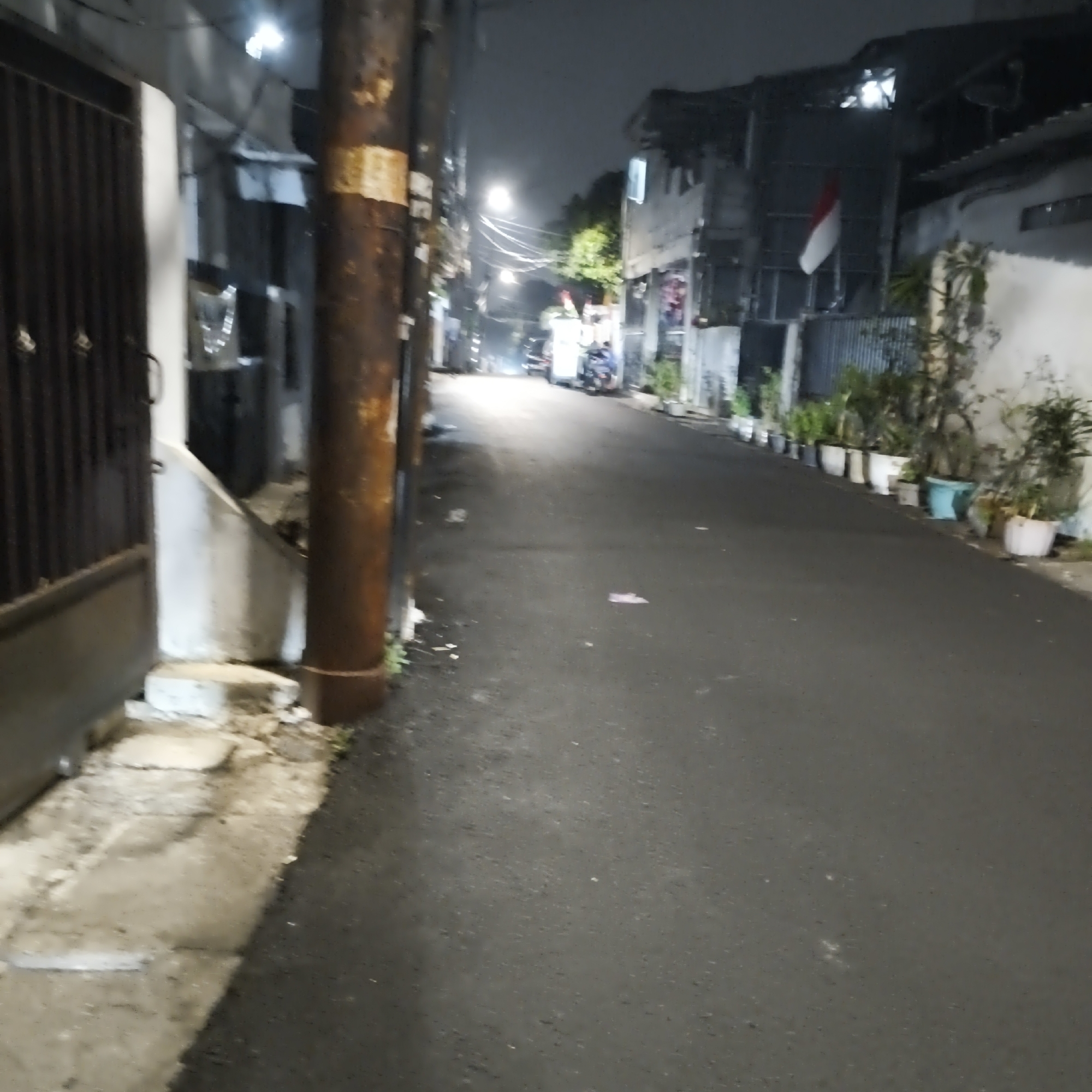 The image depicts a street scene at night in South Jakarta, Indonesia. The street is paved and appears to be a residential or commercial area with buildings lining one side. There is a prominent utility pole in the foreground with a rusted texture and some yellow and white banding. On the left side of the image, a dark-colored metal gate is visible, with a white-painted concrete structure next to it. The right side of the street is lined with potted plants. In the background, streetlights illuminate the road, and there are a few motorbikes visible. A red and white flag, likely the Indonesian flag, is partially visible on one of the buildings. The overall atmosphere is dimly lit, consistent with nighttime.