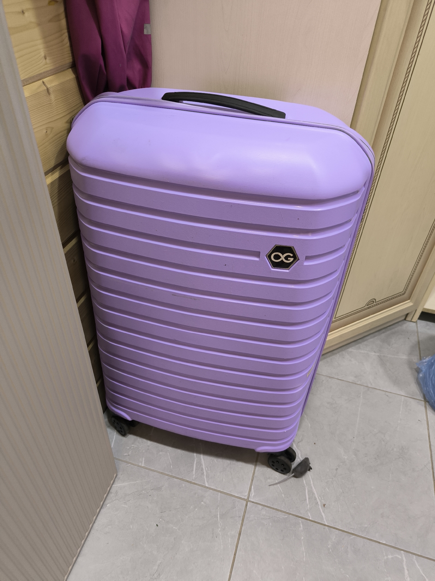 A light purple, hardshell suitcase stands upright on a grey tiled floor. The suitcase has a textured surface with horizontal ridges and a black handle on top. A hexagonal emblem with the letters "OG" in gold is affixed to the front of the suitcase. The suitcase is positioned in a corner, with a light wood-paneled wall on its left and a cream-colored cabinet with decorative molding on its right. A piece of dark pink fabric, possibly clothing, is draped over the wood-paneled wall. The floor tiles are large and rectangular with a marble-like pattern. A small, grey toy mouse is lying on the floor near the wheels of the suitcase. The scene appears to be an indoor residential space. There is no information in the image to suggest a specific location within the city of Октябрьский, Russia.