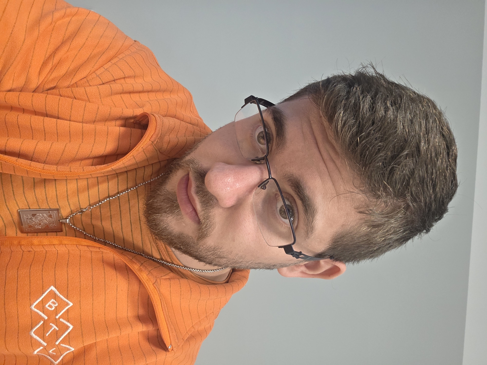 A man wearing an orange, striped hoodie and black-framed glasses is positioned in front of a light gray wall. He has short brown hair and a beard. A silver chain necklace with a pendant is visible. The hoodie has a white geometric logo on the left side. The image's orientation is rotated. The location is unknown and cannot be determined.
