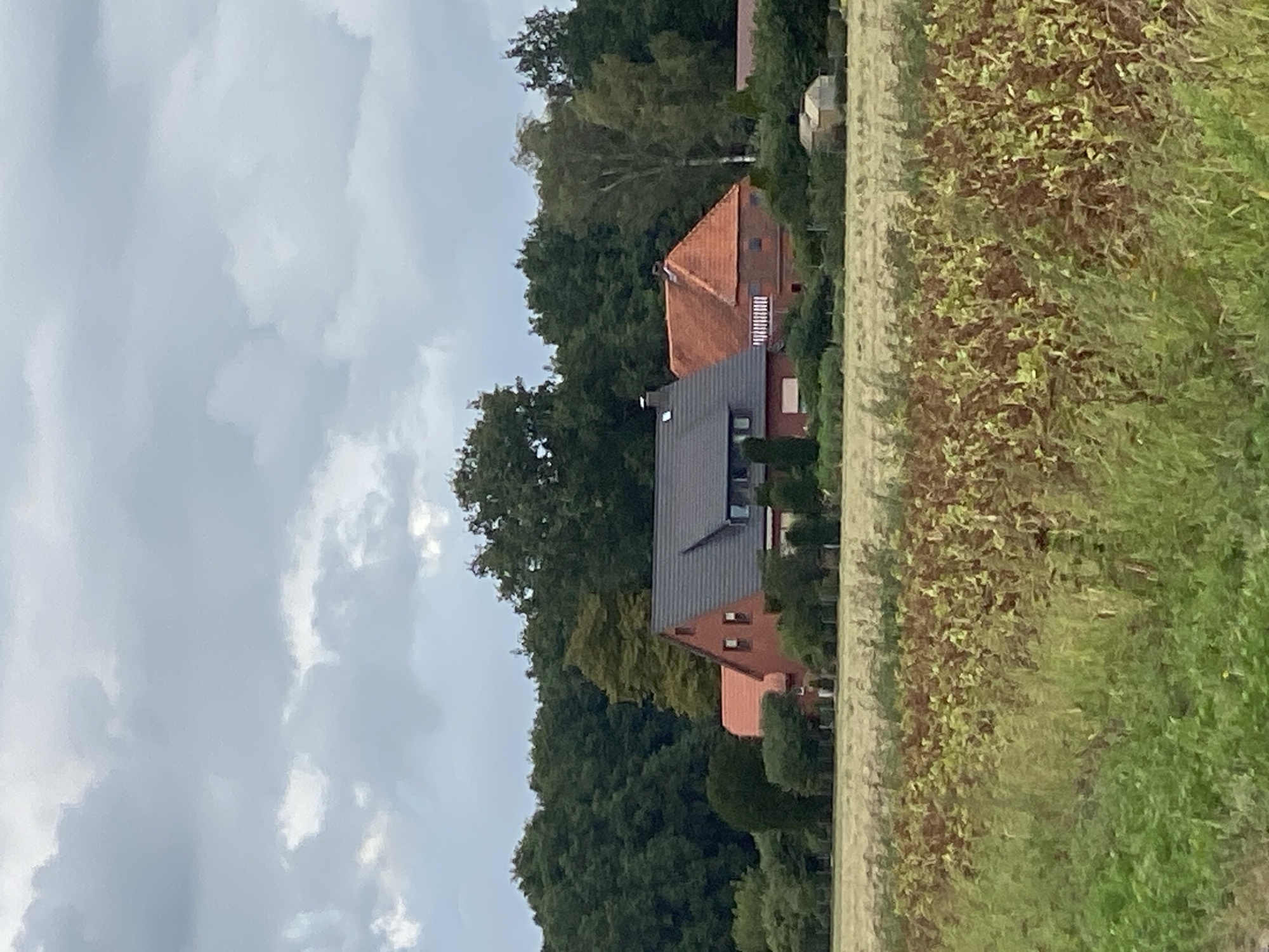 A cloudy, overcast day is captured in this vertical, medium-distance shot of a rural scene. In the foreground, a field of dry, brown vegetation with some green growth dominates the bottom half of the frame. Beyond this, a line of trees forms a dense, dark green backdrop. In front of the trees, a large, red-brick house with a dark gray roof and several windows is visible. The house has multiple sections and a prominent chimney. To the right of the main house, a smaller red-brick structure with a red tiled roof is attached. Further to the right, a small, light-colored shed or outbuilding is situated near the edge of the trees. The sky is filled with heavy, gray clouds. The scene appears to be located in or around the village of Schwarme, Germany, based on the architectural style and landscape.