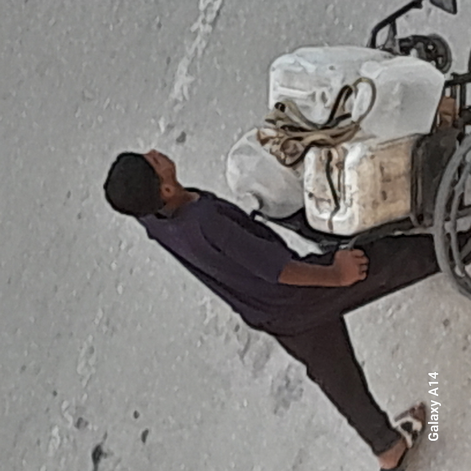 A person wearing a dark blue long-sleeved shirt and dark pants, with a black cap on their head, pushes a cart loaded with items. The cart appears to be a two-wheeled vehicle with a white cover over some of the contents, and straps securing them. The person is barefoot except for a sandal on their left foot. The background consists of a textured, greyish surface, likely a road or paved area. A watermark reading "Galaxy A14" is visible in the bottom left corner, suggesting the image was taken with that device. The overall scene depicts a person engaged in moving goods via a cart in what is described as the city of Khan Yunis, Palestinian Territories.