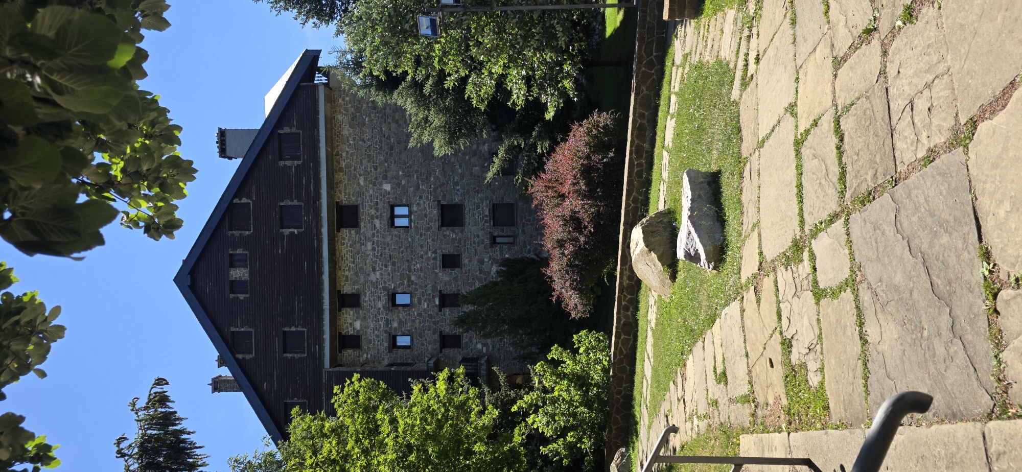 The image depicts a stone building with a dark wooden roof, surrounded by lush green trees and foliage. In the foreground, a pathway made of large, irregular stone slabs winds through a grassy area. Two large rocks are placed on the grass near the pathway. The building, which appears to be a hotel or guesthouse, has numerous small windows and is nestled amongst the trees. The sky is a clear, vibrant blue. The scene is tranquil and picturesque, suggesting a natural setting. This image was taken in Sallent de Gállego, Spain, a municipality in the province of Huesca, Aragon.