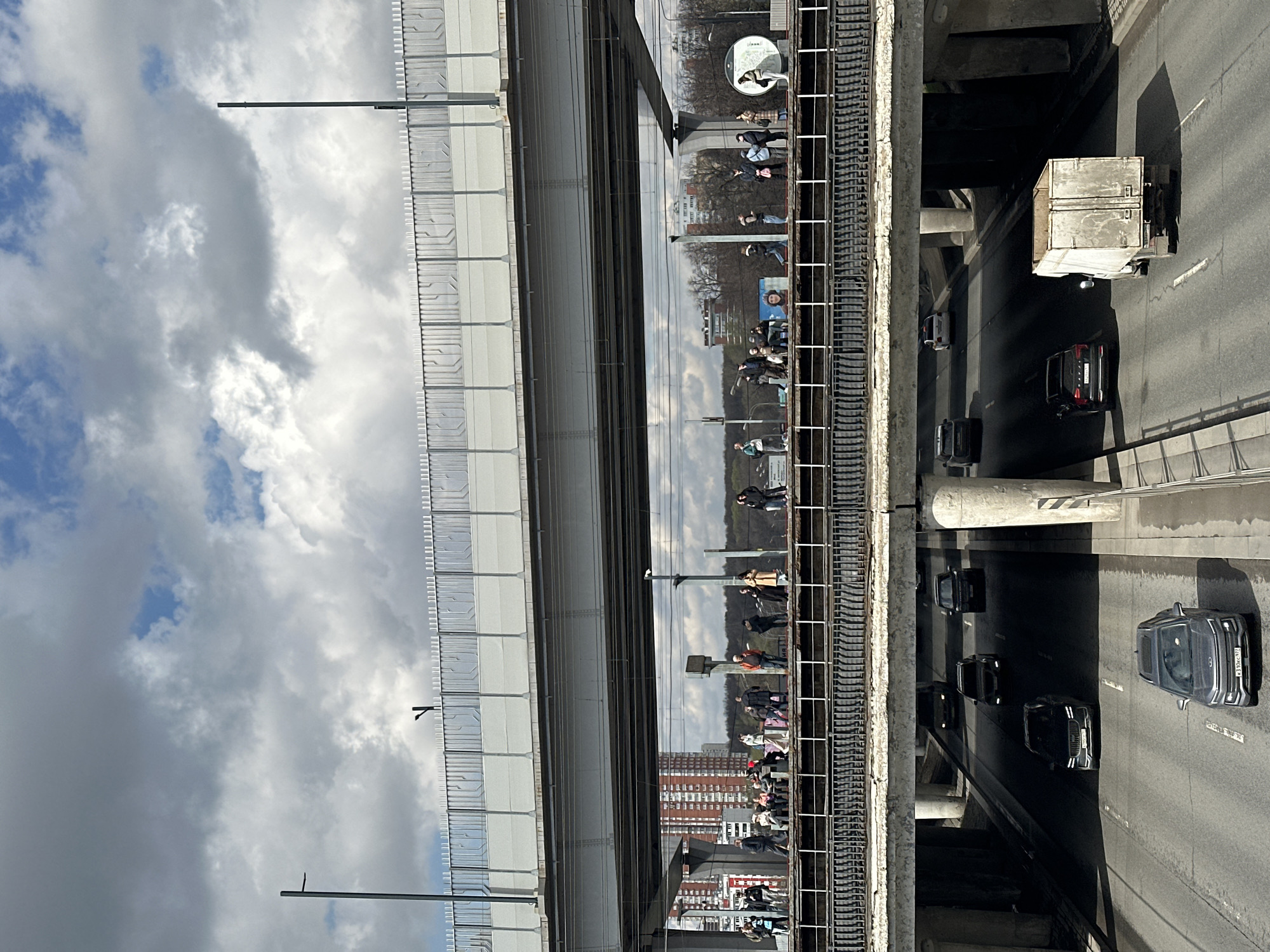 The image presents a multi-layered urban environment captured from an elevated perspective, showing infrastructure, people, and vehicles under a partially cloudy sky. The scene is illuminated by natural daylight, casting distinct shadows.
In the upper portion, an elevated structure features a bright, light-colored facade, possibly sound barriers, with a decorative railing pattern, set against a backdrop of white and grey clouds with occasional blue sky. A tall, slender pole extends vertically from this structure.
The mid-ground is dominated by a long, dark, highly reflective surface, likely a glass wall of an elevated platform or building. This surface vividly mirrors the surroundings on its opposite side, revealing numerous people standing and walking along what appears to be a walkway or platform. They are dressed in casual attire, engaged in daily activities. Reflected trees, buildings, and a circular white sign with an indistinct symbol are also visible. Below this reflective section, a track-like or textured support structure runs horizontally.
The lower section of the image shows a ground-level highway or multi-lane road. Concrete pillars supporting the elevated structures cast long, dark shadows across the asphalt. Several vehicles are present, including a dark SUV on the right lane displaying a Russian license plate "P310HC777". Another dark car is visible further down, and a light-colored truck with a grey container travels in the left lane. White road markings are visible on the asphalt surface.
The image conveys a sense of a busy urban setting with complex, vertically integrated transportation and pedestrian areas.