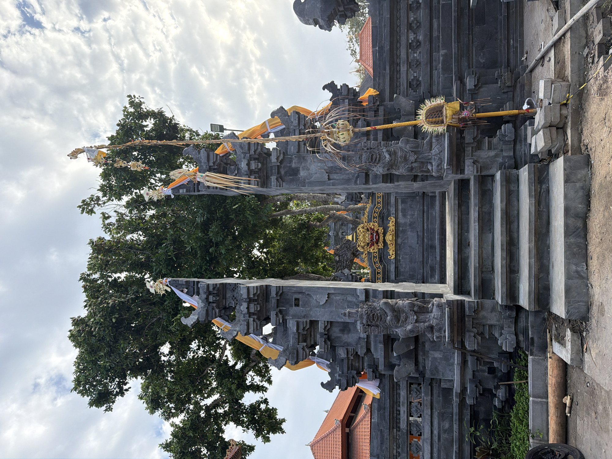 The image shows a stone temple entrance in Kuta Selatan, Indonesia, adorned with orange and white fabric banners and traditional decorations. A large tree with dense green foliage stands behind the structure. The temple features carved stone figures flanking the entryway and a decorative golden facade above the stairs. The area in front of the temple appears to be under construction, with stacked bricks, wooden logs, and a portion of a wheel visible. The sky above is partly cloudy.