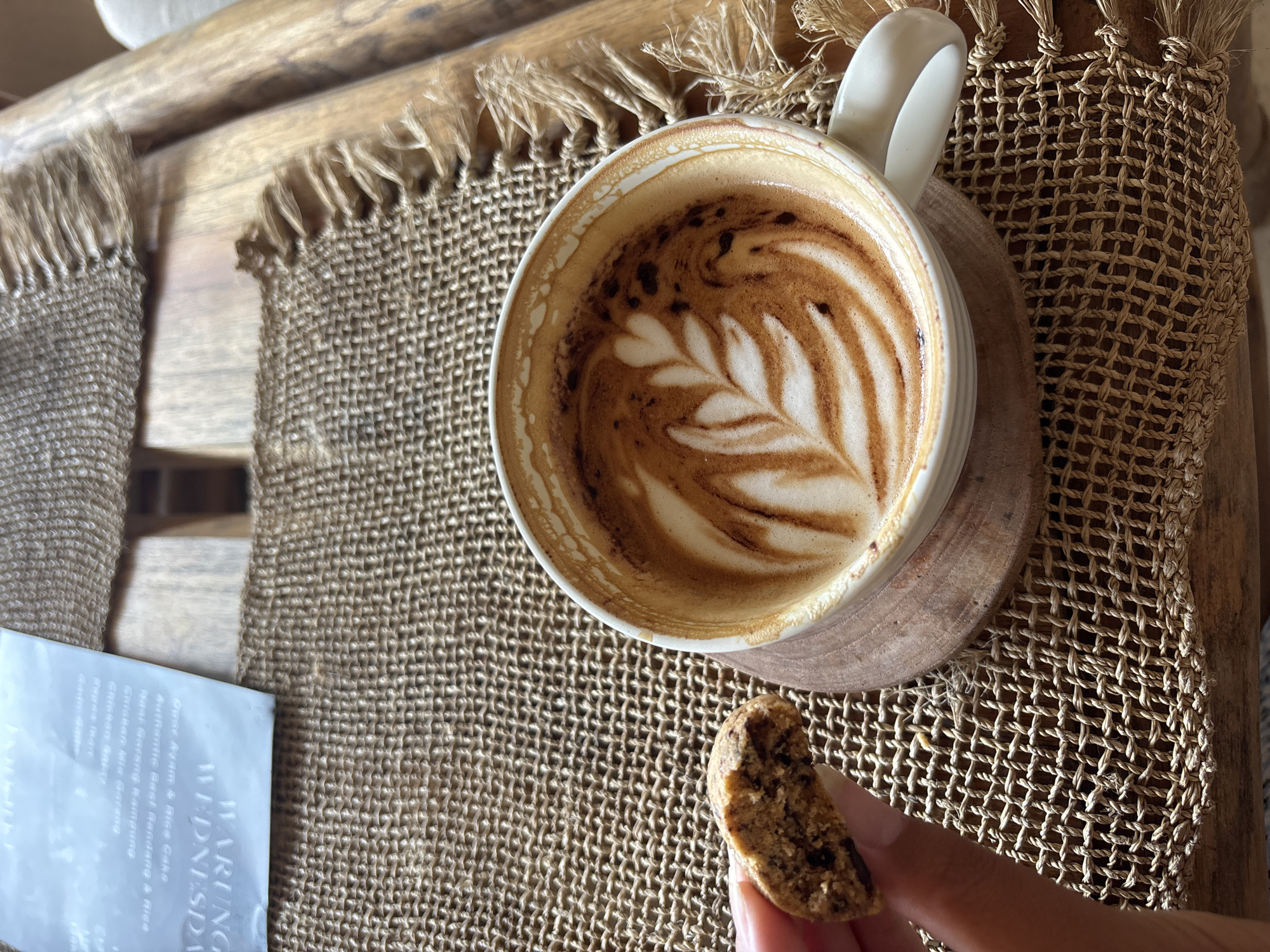 The scene is a still life of a coffee mug, cookie, and menu resting on a woven placemat atop a wooden surface. The coffee, appearing as a latte or cappuccino, features latte art in the form of a leaf. A partially eaten cookie is held in a hand. The menu text includes "WARUNG WEDNESDAY" along with a list of dishes (e.g. Ayam & Rice Cake, Rendang). Based on the menu content, the Warung Wednesday event appears to be located in Kuta Selatan, Indonesia.